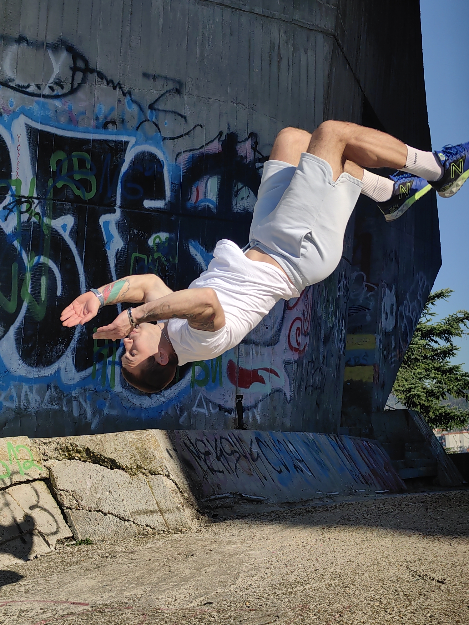 The image shows a man performing a backflip near a concrete structure covered in graffiti. He is wearing a white t-shirt, light gray shorts, white socks, and blue athletic shoes. The concrete structure appears to be part of an underpass or bridge, with a tree visible in the background. The ground in the foreground is a mix of concrete and gravel. The scene is set under a clear blue sky. Based on the graffiti style, specific tags present, and the architectural style of the underpass which is common in older parts of the city, this location is likely an underpass in the city of Varna, Bulgaria.