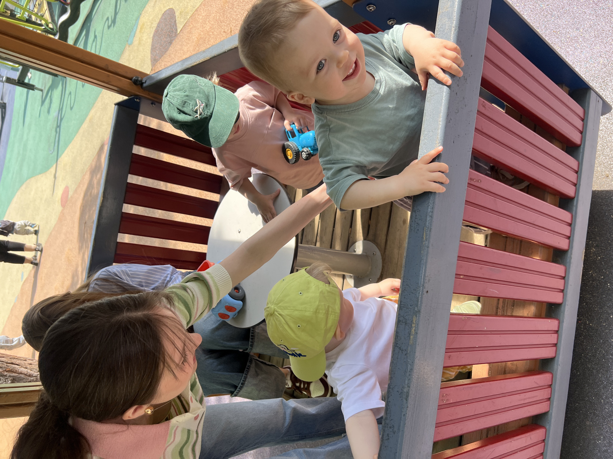 The photograph depicts a group of children playing on a playground structure. One child, wearing a light green shirt, is smiling and looking directly at the camera while grasping onto the grey railing of the play structure. Another child in a peach-colored shirt and a green baseball cap with an emblem stands nearby, holding a blue toy tractor. Below them, another child with a yellow baseball cap is also part of the group. An adult woman is present, seen from a downward angle, wearing jeans and a striped sweater; her left arm is outstretched towards the children. The structure itself is multi-colored, with grey framing and red horizontal slats. Parts of the wider playground area are visible, as is a small portion of a mural. The exact location in Moscow, Russia, cannot be determined from the visual information provided.