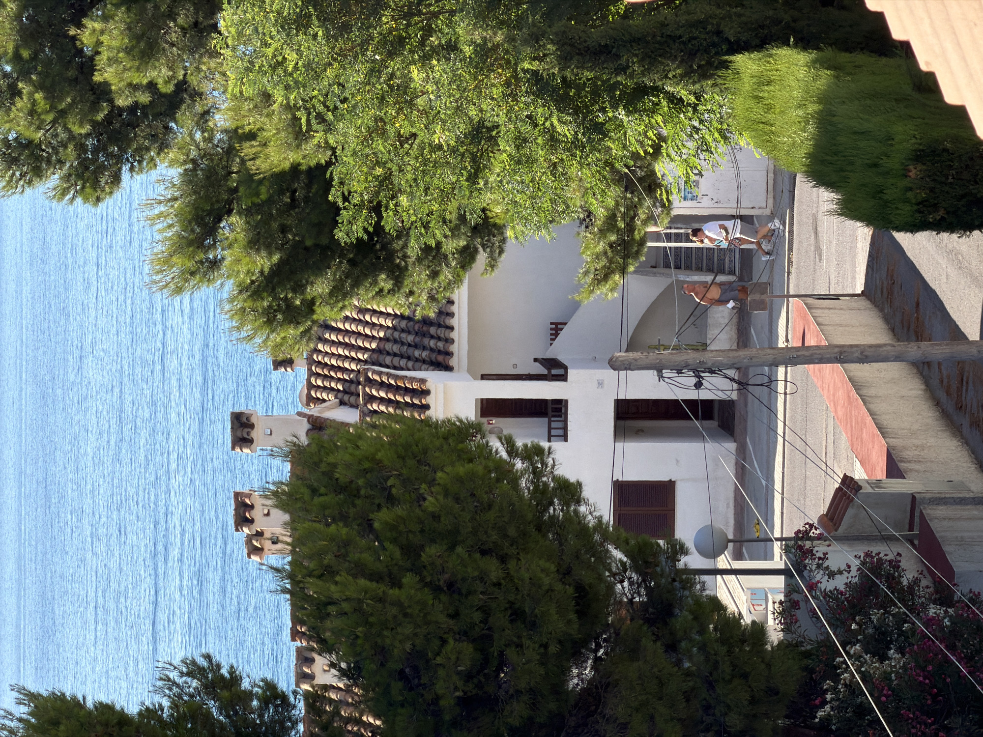 The image shows a view of the Mediterranean Sea from a residential area in Alcalà de Xivert, Spain, specifically from a vantage point looking over rooftops and trees towards the water. A white building with a terracotta tile roof and decorative chimneys is prominent. In front of the building, two men are walking. One man, shirtless and wearing grey shorts, is walking from left to right. Behind him and slightly to the right, another man in a white t-shirt and grey shorts is walking in the same direction. A lush green pine tree is in the foreground on the left, and a dense green hedge is on the right. The sea is visible in the background, a clear blue expanse. Electrical wires and a lamppost are also present in the scene.