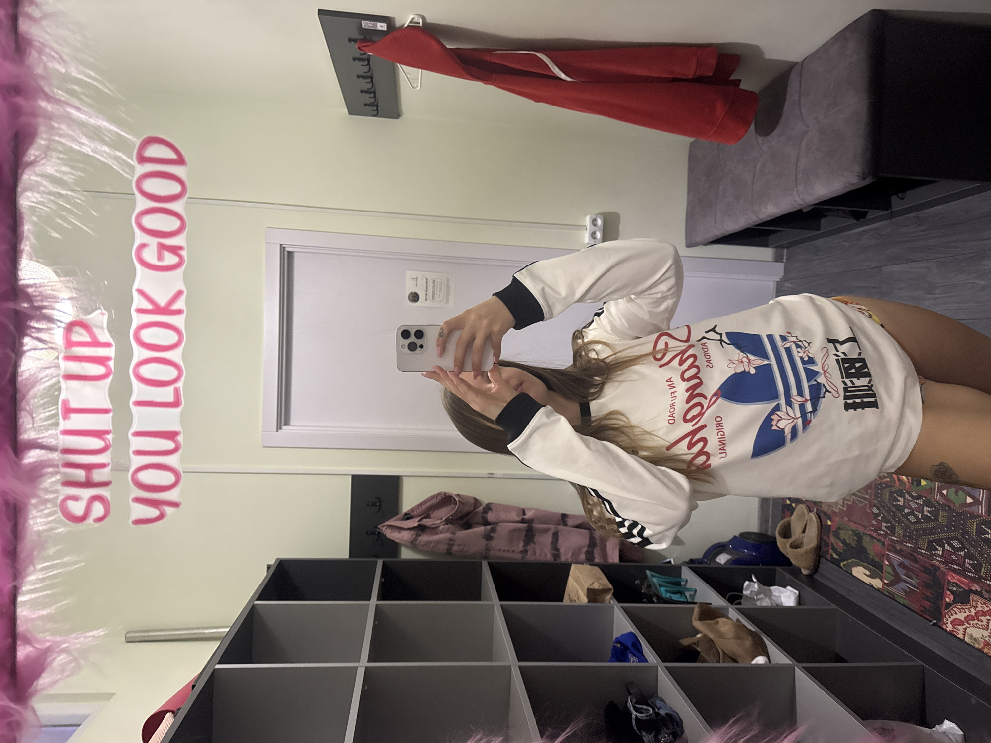 A young woman takes a mirror selfie in a fitting room. The fitting room features a pale green wall with a pink, furry border and a sign that says "Shut up You Look Good". In the reflection, a red jacket hangs on a coat rack above a door. A gray bench and dark gray flooring appear on the upper right side of the image. Below the mirror, there is a gray shelving unit with various objects, including shoes, bags, and clothing, filling the compartments. The location is likely a clothing store fitting room. The city of Batumi, Georgia cannot be confirmed.