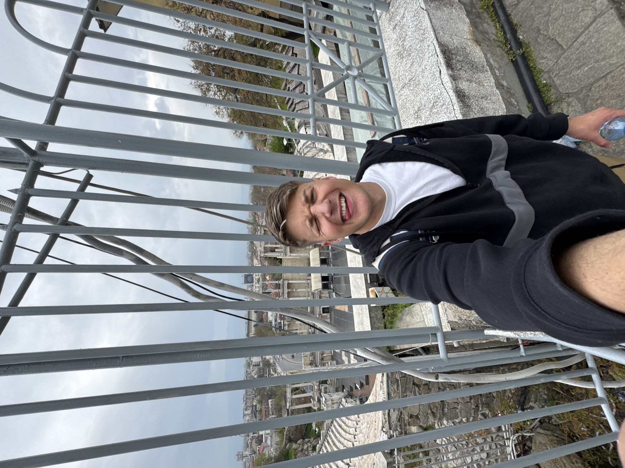 A selfie shows a man standing behind a grey metal railing, with a cityscape visible in the background. The man is wearing a white t-shirt under a black jacket with grey stripes, and he is holding a water bottle in his right hand. The cityscape below includes streets, buildings, and architectural ruins. The image suggests an elevated viewpoint, possibly from a tower or tall structure. Given the architectural features, the location could be Plovdiv, Bulgaria, specifically the remains of the Ancient Theatre of Philippopolis.