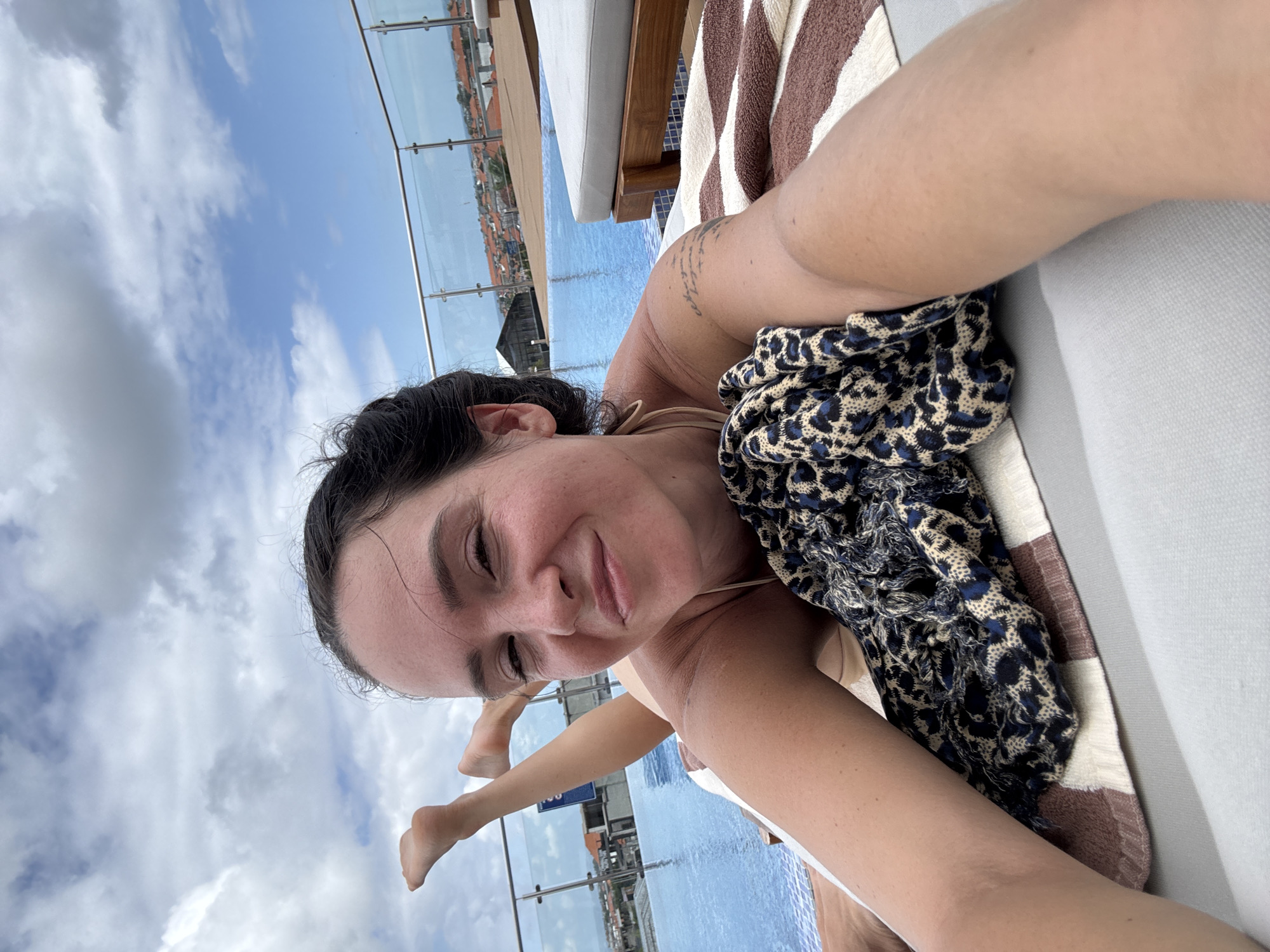 A woman with dark hair, wearing a beige bikini top, reclines on a chaise lounge by an infinity pool. Her legs are propped up, one bent and the other extended upwards. A patterned scarf is draped over her midsection. The background shows the blue water of the pool, a railing overlooking a city with red-tiled roofs, and a cloudy sky. The scene appears to be on a rooftop in North Kuta, Indonesia.