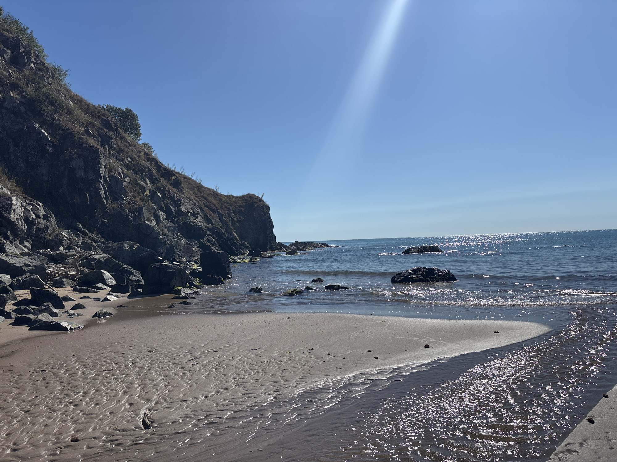 This image depicts a natural seascape on a clear, sunny day, likely in Rezovo, Bulgaria. The foreground features a sandy beach with intricate patterns of water drainage, suggesting the tide has recently receded. A narrow stream of water flows across the sand, reflecting the bright sunlight. To the left, a rocky cliff face rises steeply, covered with sparse vegetation and small trees. The cliff is interspersed with large boulders and smaller rocks, some of which are at the edge of the water. The sea stretches out to the horizon, its surface shimmering with sunlight. Several dark rocks or small islets are visible in the water near the shore. The sky is a brilliant, cloudless blue, with a strong beam of sunlight cutting diagonally from the upper right corner of the frame, creating a lens flare effect. The overall scene is serene and unpopulated.