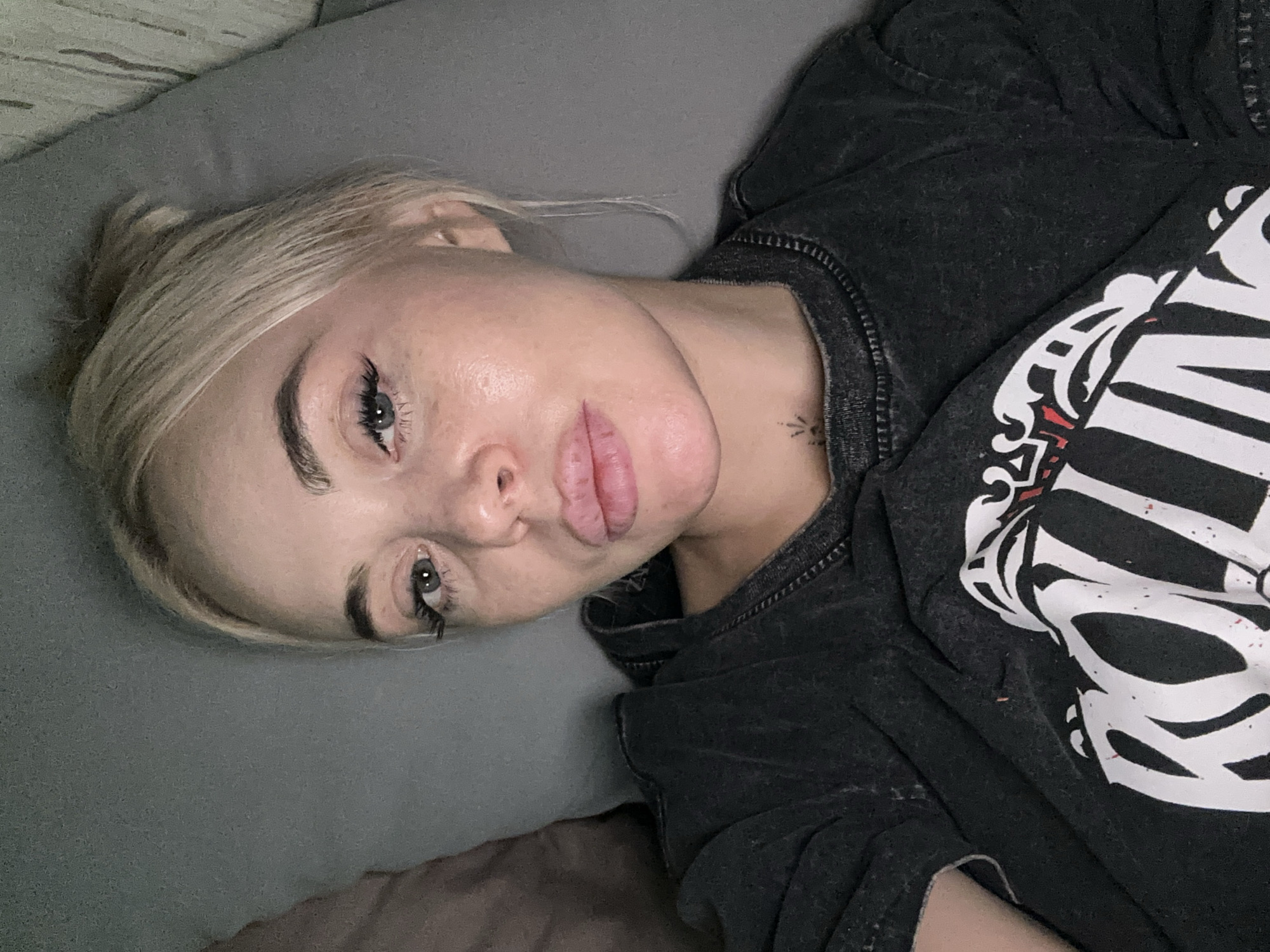 A fair-skinned young woman with blonde hair pulled back in a bun is lying down on a grey cushion. She is wearing a dark grey Rolling Stones band t-shirt. The image is a close-up, focusing on her face and upper body. A tattoo is visible on her neck. The location cannot be determined from the image.