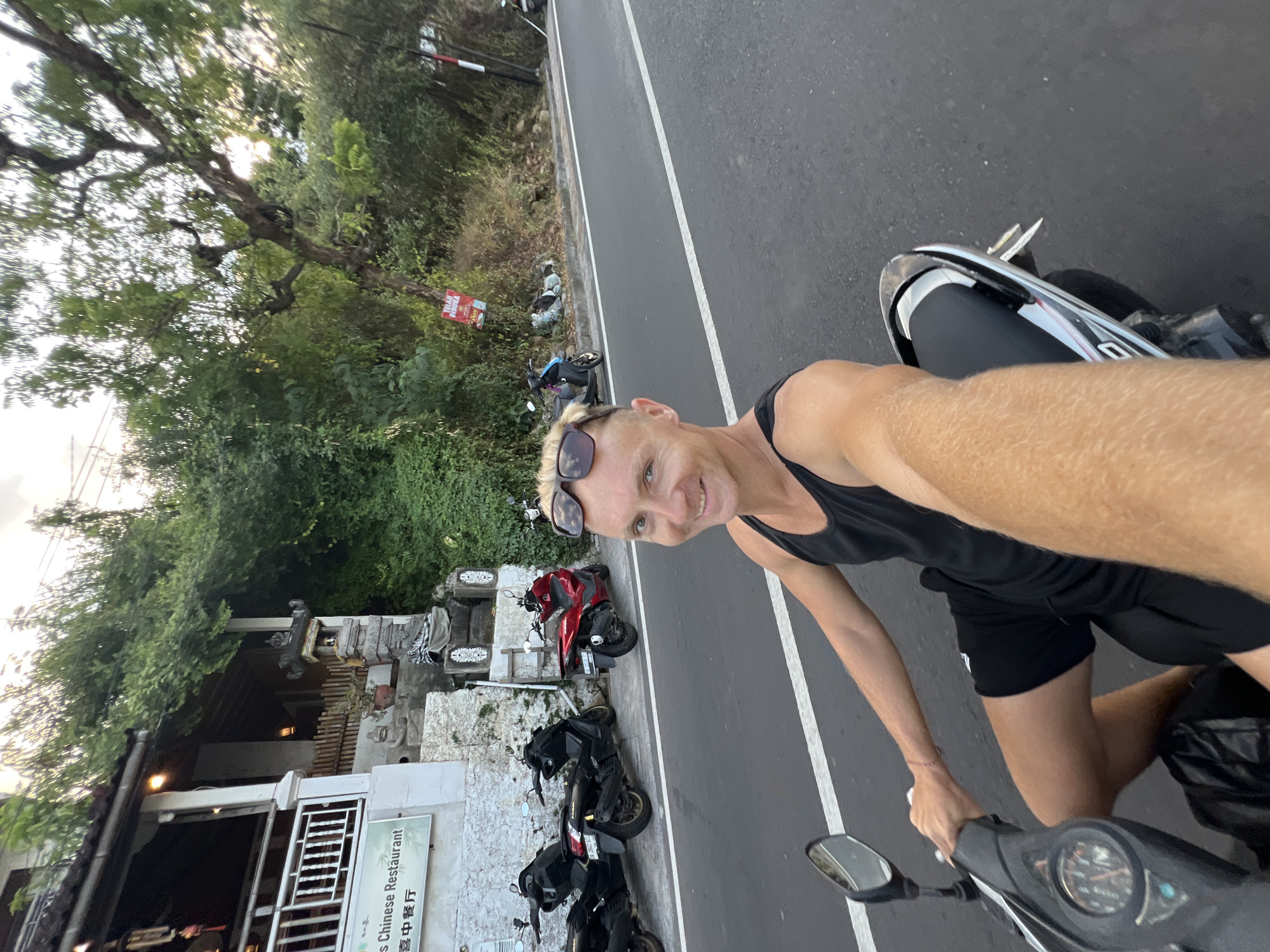 A Caucasian male with blonde hair, wearing sunglasses, a black tank top, and black shorts, is riding a scooter on a paved road in what appears to be a tropical environment. He is holding the handlebars with his left hand and has his right arm extended, holding a phone to take a selfie. The scooter is black and silver with a partially visible instrument panel. In the background, there are several other scooters parked along the side of the road. A building with a white facade, a balcony, and signage that reads "s Chinese Restaurant" is visible on the left. Dense green foliage and trees line the road and the building. A red sign with white text is attached to a tree on the right side of the road. The lighting suggests it is either early morning or late afternoon. The location is identified as Abang, Indonesia.