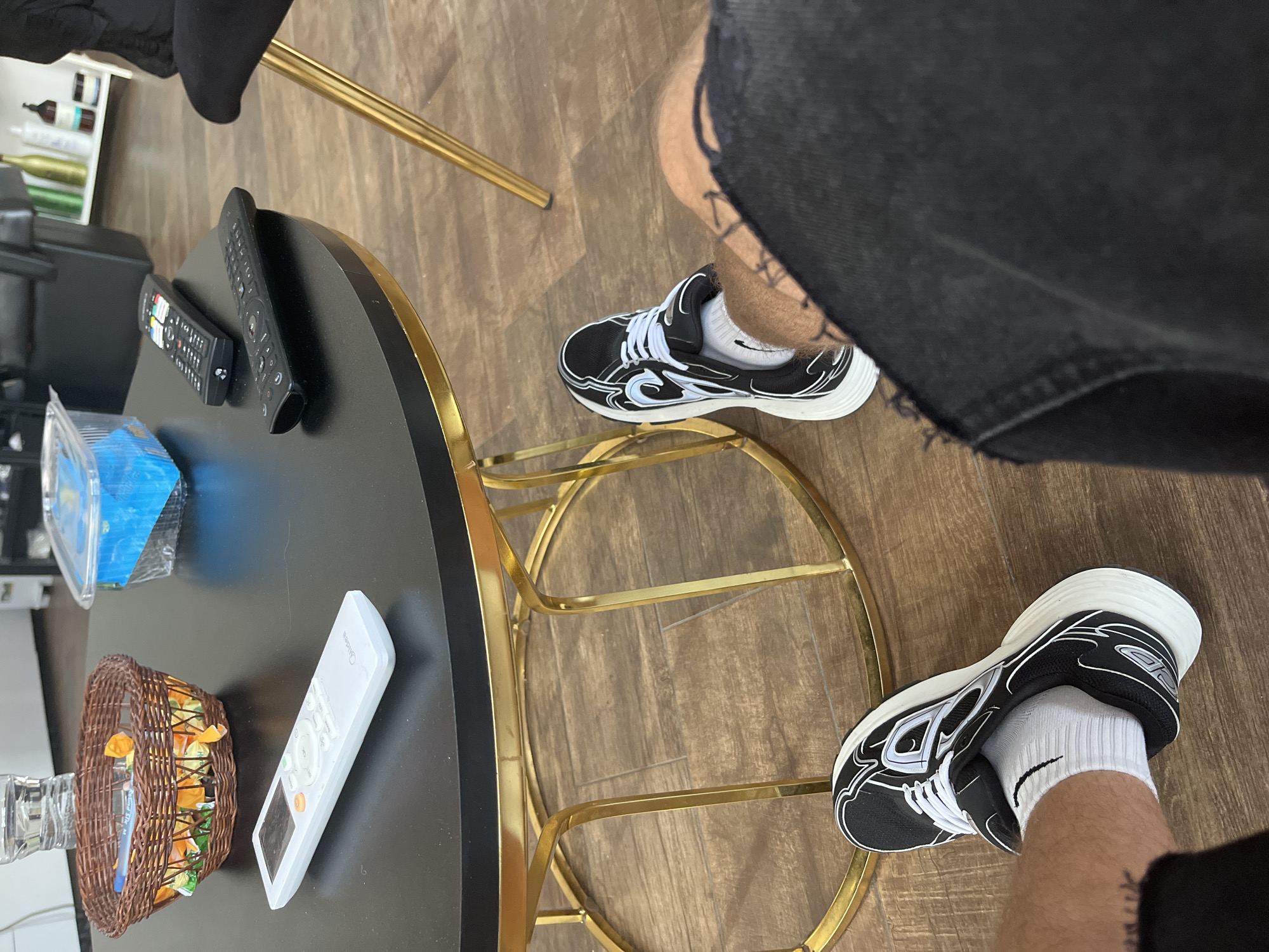 An indoor, first-person perspective photograph depicts the legs of an individual seated in what appears to be a waiting area or lounge. The person's legs are clad in white socks with a black stripe and black athletic shoes featuring white soles and light blue/white design elements. Black fabric, possibly shorts or distressed trousers with frayed edges, covers the upper legs.
In the foreground, a round black coffee table with a gold-colored metal base is visible. On the tabletop, from left to right, are: a brown wicker basket containing individually wrapped candies in yellow/orange wrappers; a transparent plastic water bottle; a clear plastic container with blue contents or a blue lid; a white remote control branded "Midea" with orange and grey buttons; and two black remote controls, one displaying buttons for streaming services like Netflix and YouTube.
The floor is covered with light brown wood-look flooring. In the blurred background, to the right, shelving units holding various bottles (possibly hair or beauty products) are discernible, suggesting a salon or similar commercial establishment. A black chair with a gold leg, matching the table's design, is also partially visible. The image contains no visual information to confirm the specific location as Zlatograd, Bulgaria.