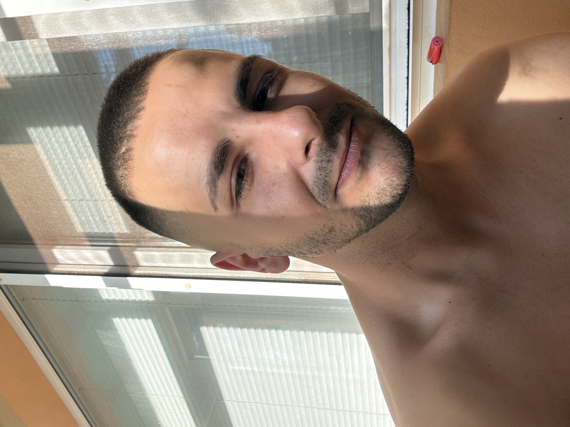 The image shows a close-up, outdoor portrait of a young man with a buzz cut and stubble. He is shirtless, with his bare shoulders and upper chest visible. The man is looking slightly off-camera with a relaxed expression. Sunlight streams in from the right, casting shadows across his face and the background. The background features a window with blinds and a warm-toned wall. A small, red object, possibly a lighter, rests on the windowsill on the right side of the frame. The scene appears to be taken on a balcony or near a window of a building in Lovech, Bulgaria, judging by the architectural style of the window and the quality of light.