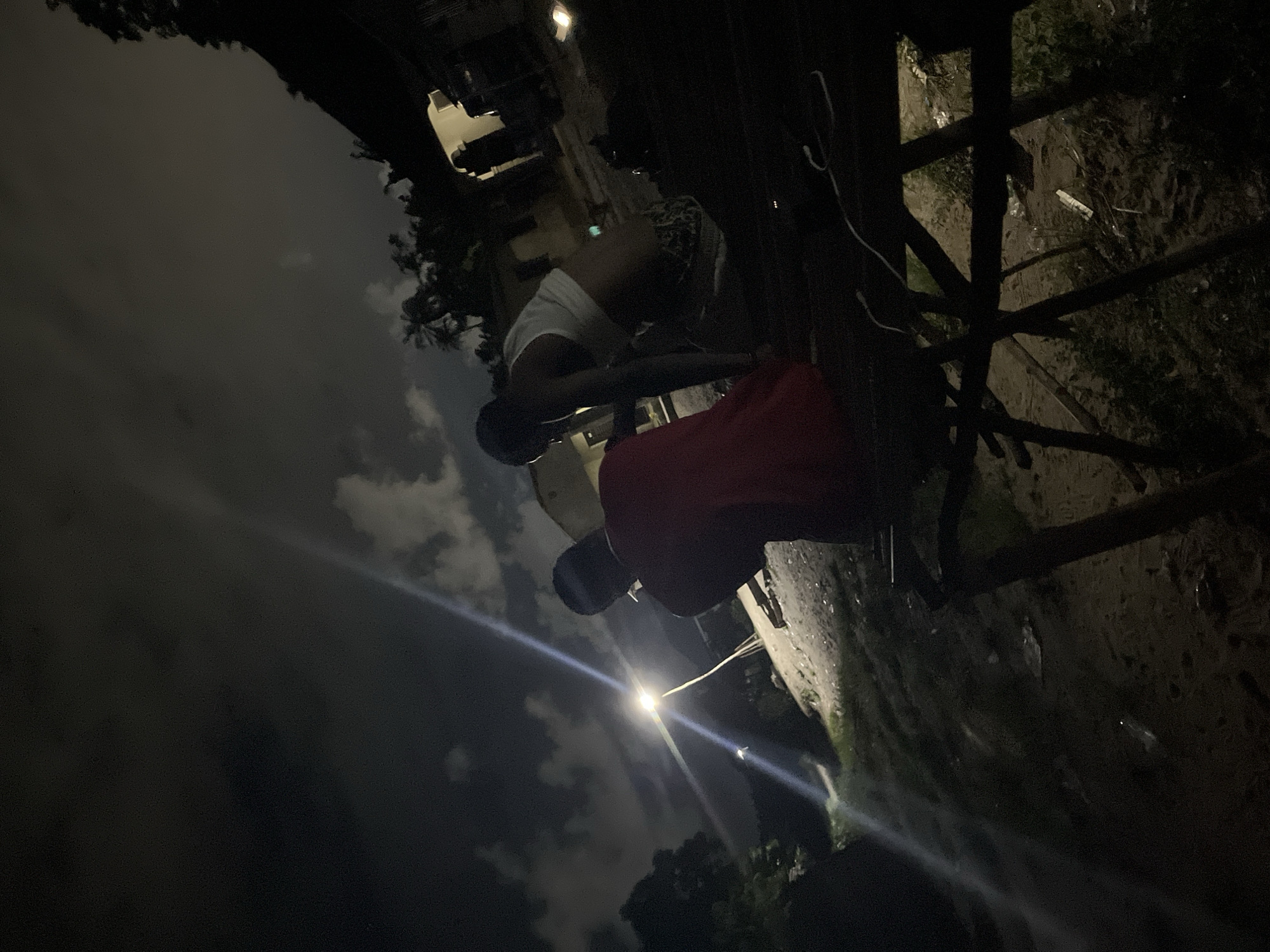 Two individuals are present outdoors. One individual is wearing a white shirt, while the other is wearing a red shirt. The location appears to be a city street at night, based on the presence of street lights and buildings. A structure resembling a fence or railing is visible. The image quality is somewhat dark.
