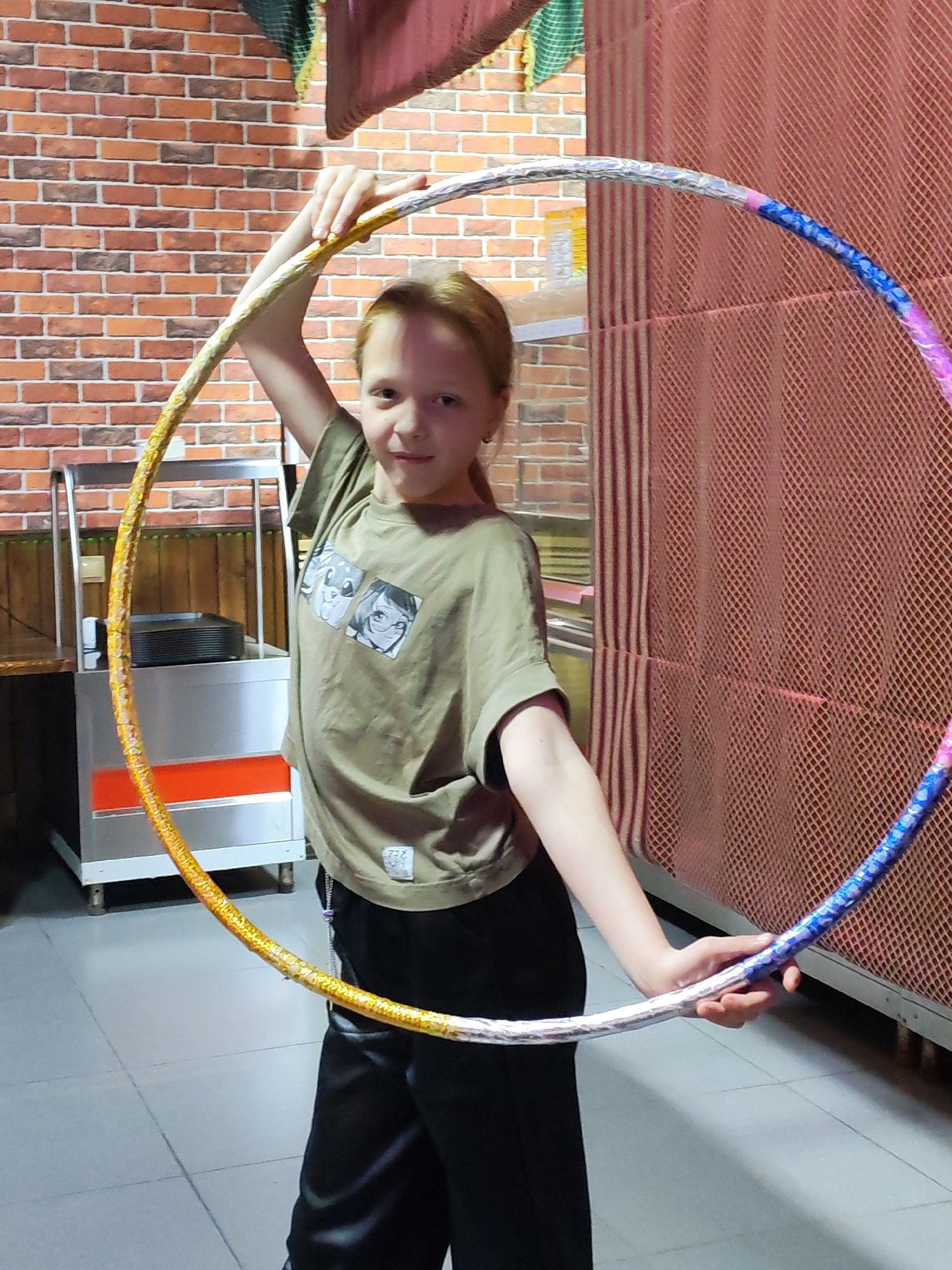 An indoor, full-body portrait captures a young girl, possibly in Chaikovsky, Russia, holding a large, colorful hula hoop. The girl, who appears to be pre-teen with light reddish-brown hair, stands facing the camera with a neutral, slightly smiling expression. She wears a relaxed olive-green t-shirt featuring two anime-style graphic prints on the front and black pants. Her hands are positioned, one overhead and one lower, to hold the hula hoop in front of her, creating a frame around her upper body. The hula hoop is visibly decorated with shiny, patterned tape; one large section appears golden or orange, while the opposite side features silver, blue, and purple segments.
The background features a rustic brick wall on the left, next to a stainless steel service cart with several black trays stacked on it. To the right, a reddish-brown, textured mesh screen or partition extends upwards, partially obscuring a solid surface behind it. The floor is made of light-colored tiles. The lighting suggests an indoor setting, possibly a public or commercial space like a cafe, restaurant, or recreation area. Small, illegible white text or a logo is faintly visible on the lower front of her t-shirt, beneath the graphic prints. The overall scene suggests a casual moment, possibly posing with her hula hoop during a break or after an activity.