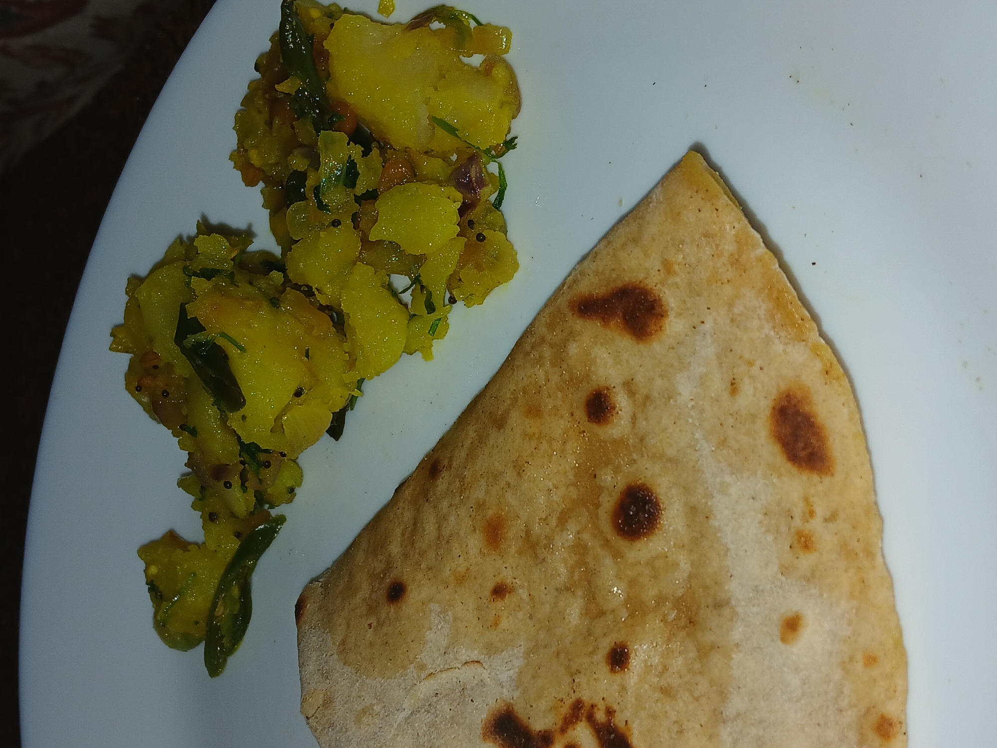 The image shows a plate of food, consisting of a flatbread (chapati or roti) and a yellow-colored vegetable dish, likely potato (aloo) sabzi. The sabzi appears to have green chilies and other spices mixed in. The food is presented on a white plate. Without additional context or landmarks, pinpointing the location within Bengaluru is not possible. The image offers no insights into people, actions, or interactions.