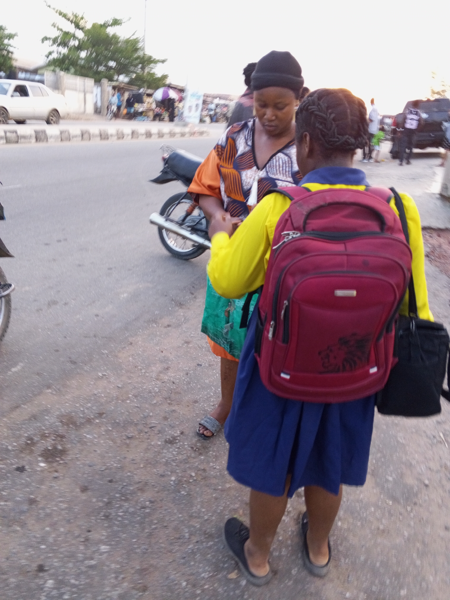 On a street in Zango-ndaji, Nigeria, a woman in a patterned orange and black top and black hat stands next to a young person. The young person wears a yellow shirt, blue skirt, black shoes, and carries a red backpack and black bag. They appear to be interacting. A motorcycle is parked nearby. The street is paved and there are other people and vehicles in the background. The sky is bright.