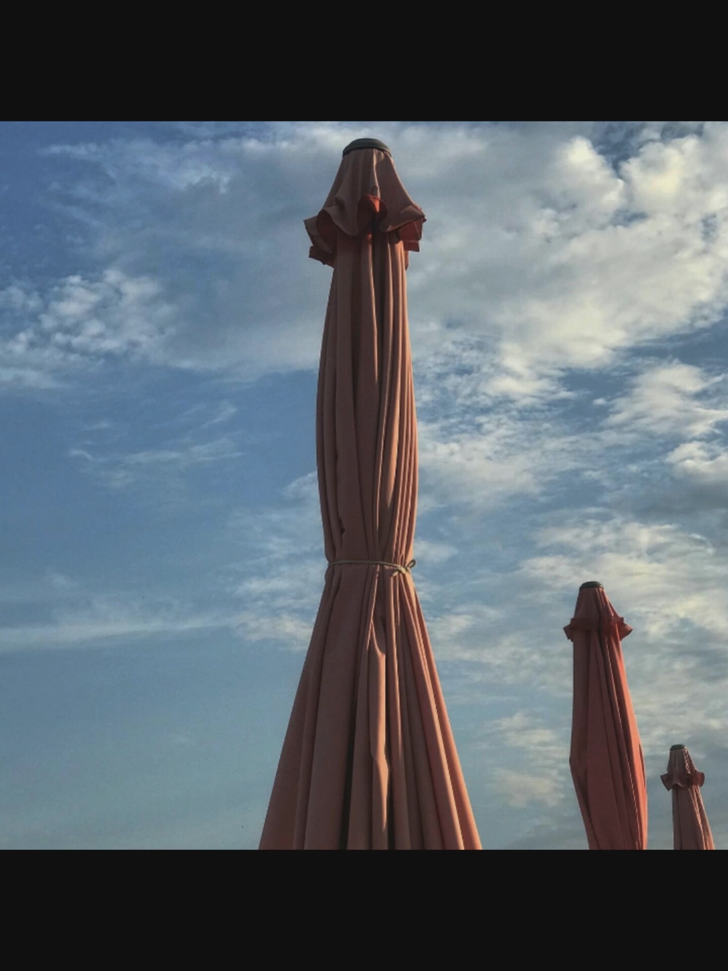 The image depicts three closed beach umbrellas against a partly cloudy blue sky. The umbrellas are a muted pinkish-red color and are closed, with their fabric neatly folded and secured with ties. The largest umbrella is in the foreground, taking up a significant portion of the left side of the frame. To the right and slightly behind it, two smaller umbrellas are visible, receding in size. The scene appears to be outdoors, possibly in a beach or park setting. There is no discernible location in Bucharest, Romania, or any people present in the image. The focus is on the umbrellas and the sky.