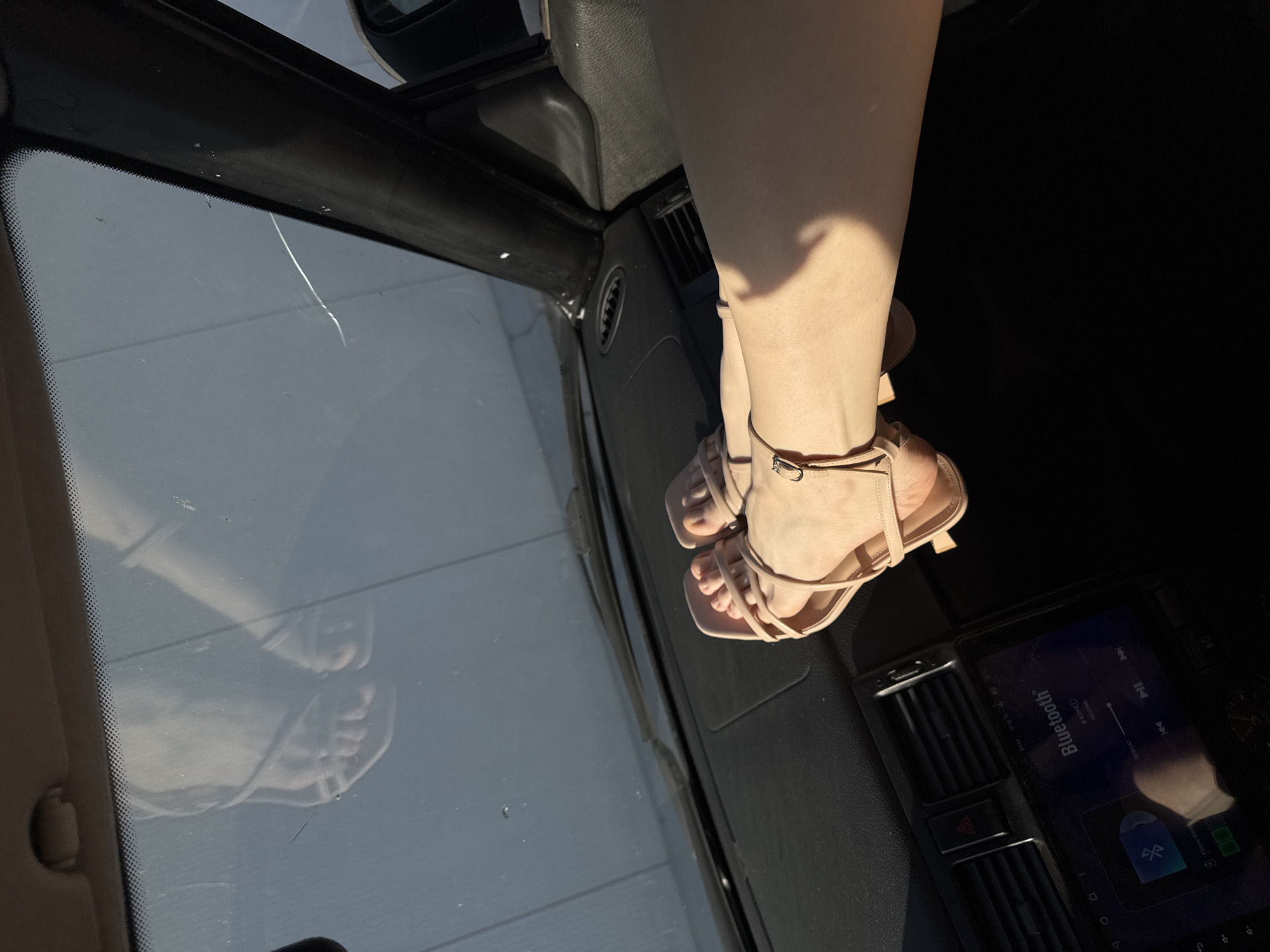 The photograph depicts the interior of a car. A person's legs and feet, clad in strappy beige heels, are positioned on the dashboard. The reflection of the feet is visible in the car's windshield. The dashboard features air vents and a digital display screen indicating a Bluetooth connection. The location is not identifiable as Perm, Russia from the available image.
