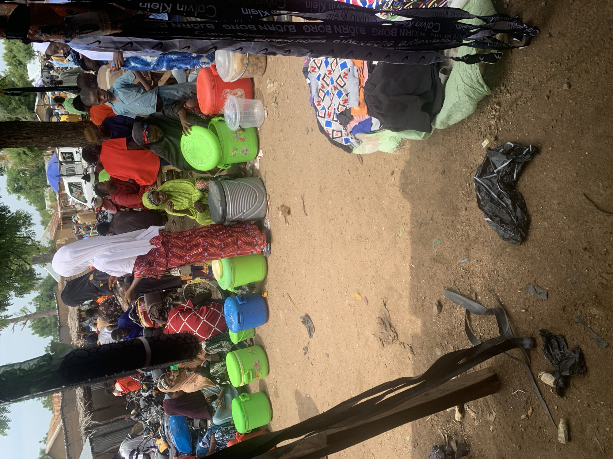 In Project, Nigeria, a bustling marketplace scene unfolds on a dusty dirt ground. The image captures a diverse group of individuals, some dressed in vibrant traditional clothing, others in more casual attire. A variety of large plastic containers, some green and others blue, are prominently displayed, suggesting they are for sale or transport. Amongst them, a woman in a red and white patterned dress and a white hijab is seen holding what appears to be a chicken. Nearby, a person in a green headscarf and dress is interacting with one of the green containers. In the background, a white truck is visible, along with several motorcycles, indicating a lively commercial hub. The overall atmosphere appears active and crowded, with numerous people milling about and engaging in various activities. The ground is uneven and scattered with debris, including plastic bags and what looks like dried corn husks, further emphasizing the outdoor market setting.
