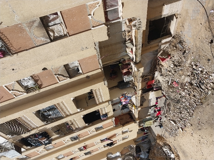 This image depicts a multi-story residential building in Khan Yunis, Palestinian Territory, exhibiting significant structural damage. The beige and reddish-brown facade features numerous balconies with white balustrades and windows, many of which are broken or have missing panes. Several windows show signs of scorching or fire damage. Laundry, including various garments, hangs from numerous balconies and clotheslines on multiple floors and at ground level, indicating continued habitation. The lower floors of the building show considerable destruction, with exposed concrete and rebar.
The ground area directly in front of the building is covered with extensive rubble and debris, consisting of broken concrete, twisted metal, and miscellaneous waste materials. A large mound of this debris extends across the foreground. On the lower right, a dark, open void suggests a collapsed or damaged ground-level entrance. On one lower balcony, multiple large black water tanks or storage containers are present. A makeshift tent or tarpaulin structure is visible on the left side at ground level amidst the debris. No individuals are clearly visible in the scene.