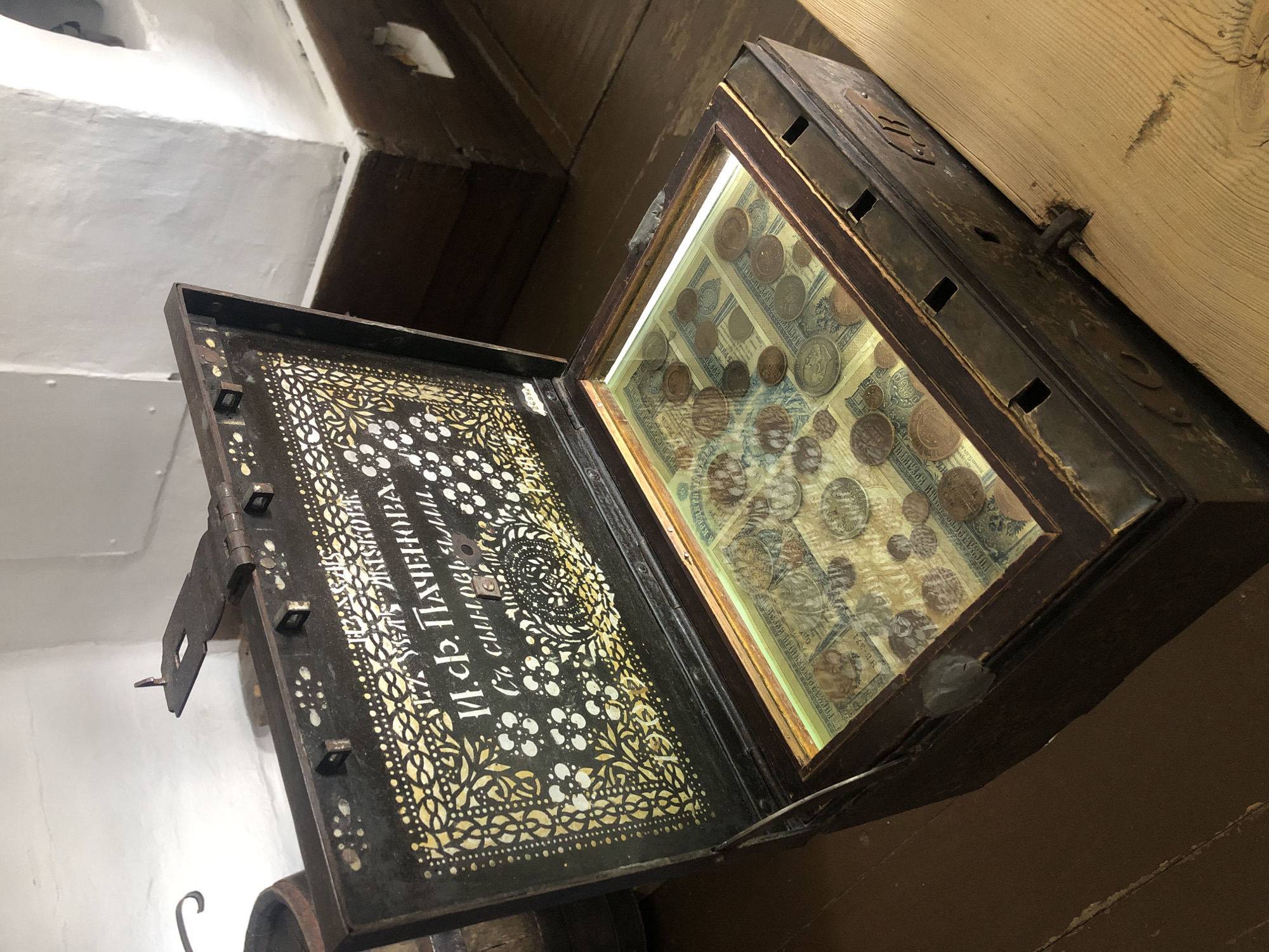 An antique wooden chest with a glass lid displays a collection of old currency and coins. The chest is open, revealing its contents. The interior of the lid is decorated with an ornate golden pattern against a dark background, featuring Cyrillic text. The collection inside includes what appear to be banknotes and various coins of different sizes and colors. The chest itself is dark wood with metal fittings. The scene is set indoors, on a wooden floor, with a white wall visible in the background, suggesting it is a historical exhibit or a private collection. This particular chest is on display at the Shushenskoye Museum of Folk Culture and Everyday Life in Shushenskoye, Russia, specifically in the exposition dedicated to Lenin's exile.