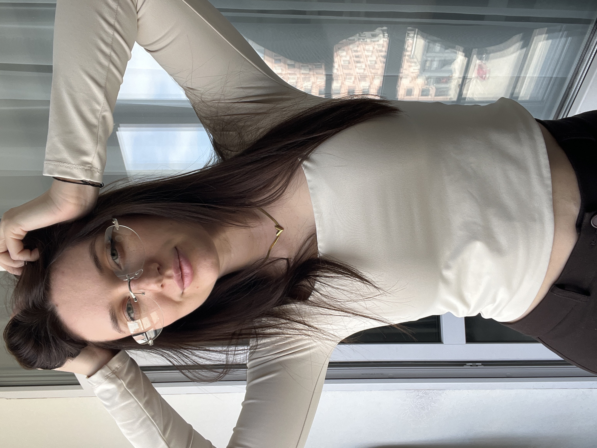 A young woman with dark hair is lying down, supporting her head with her hands. She is wearing rimless glasses, a light-colored long-sleeved shirt, and dark pants. A gold necklace is visible. The background shows a window with reflections of buildings, potentially indicating an urban environment. The location is specified as Tyumen, Russia, based on the request.