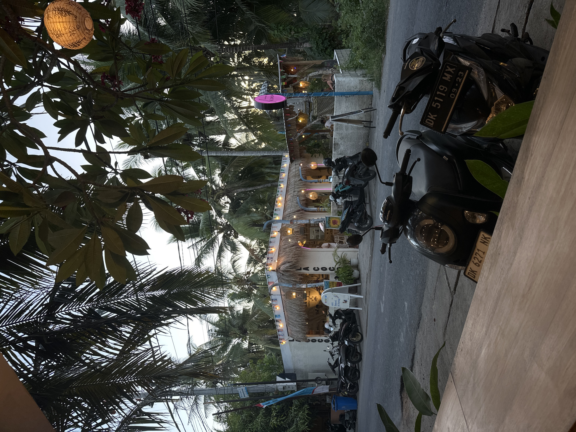 The image depicts a street scene in Nusa Penida, Indonesia. A small, open-air restaurant or café is located along the roadside, its thatched roof adorned with string lights and colorful pennants. Palm trees are seen surrounding the café. Several motorcycles are parked on the street in front of the café. A spherical paper lantern hangs from a tree branch. A sign with a pink circular logo is visible. The license plates of the motorbikes are visible, with DK 5119 MX and DK 6271 NK as examples.