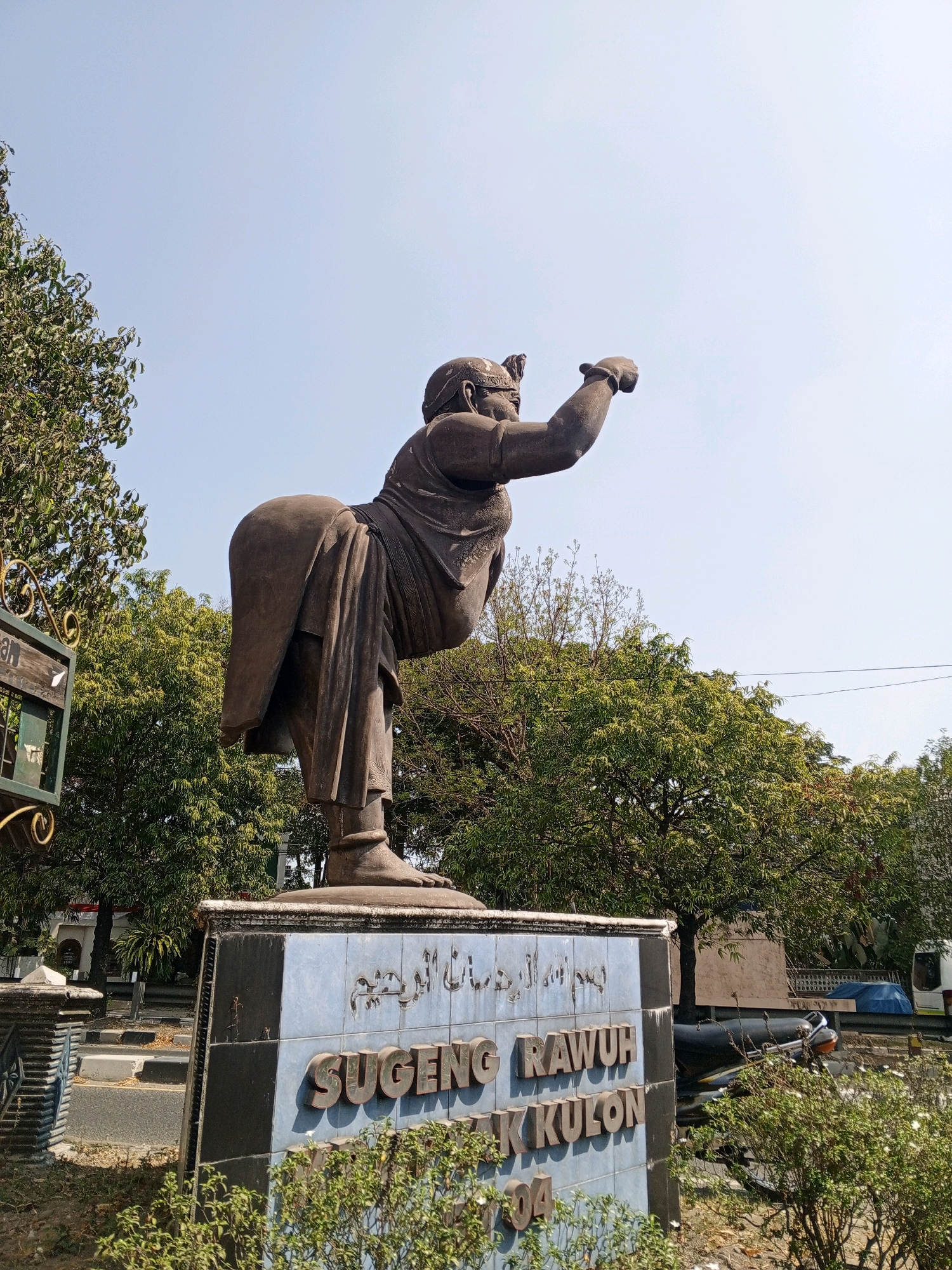 This image shows a bronze statue of a person in a dynamic pose, with one leg raised and the arm extended. The statue is on top of a pedestal with blue tiles and text that reads "SUGENG RAWUH" and "TEBAK KULON 104". The background consists of trees, a clear sky, and some urban elements like a road and buildings. The statue is located in the city of Sewon, Indonesia, likely a public space or a monument. The lighting suggests it's daytime.