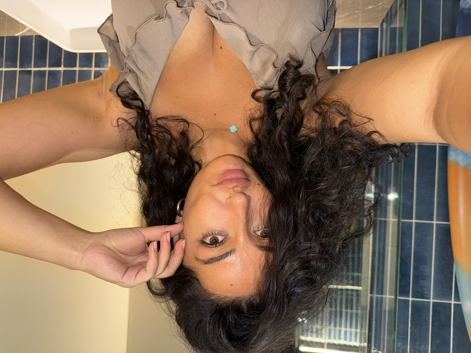 A woman with dark, curly hair and olive skin is captured in an upside-down, medium close-up self-portrait. She looks directly into the camera with a neutral expression, her brown eyes prominent. Moles are visible on her face and chest. Her lips are slightly parted, revealing a natural pink hue.
She wears a light brown or taupe top featuring ruffled edges along the neckline and a deep V-neck. A delicate necklace with a small, light blue or green pendant, possibly shaped like a flower or paw print, adorns her chest. A gold-colored hoop earring is visible in her left ear. Her left hand, with natural-colored, manicured nails, is gently placed near her temple and hair.
The background primarily consists of dark blue rectangular tiles, characteristic of a bathroom. A white fixture, likely a sink or bathtub rim, is visible in the upper left. Reflections within the tiled area suggest the presence of a mirror. Faint red marks and a lighter patch, possibly a scar, are visible on her arms.
The photograph is taken indoors, likely under artificial lighting, in Kuta Selatan, Indonesia. There is no visible text in the image.