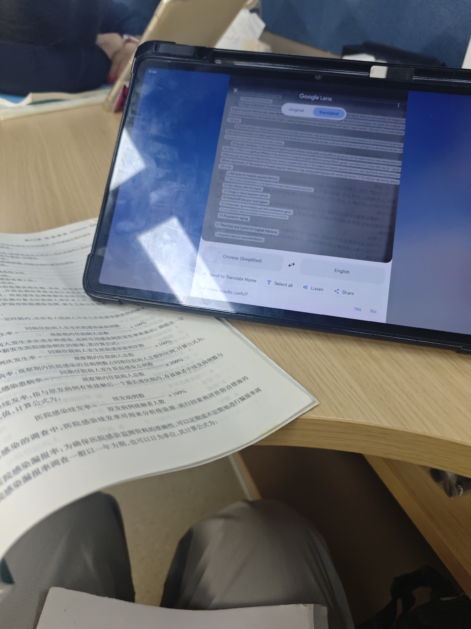 An overhead shot shows a tablet computer displaying the Google Lens app, with text translated from Chinese to English. The tablet is on a wooden desk, with a person's arm and shoulder visible on the left side of the frame, as they are partially seen sitting at the desk. Paper documents are scattered on the desk, with some partially visible in the foreground and background. The scene is indoors, with the lighting suggesting it is daytime. No specific location in the Chongqing High-tech Zone, China is identifiable from the visual information provided.