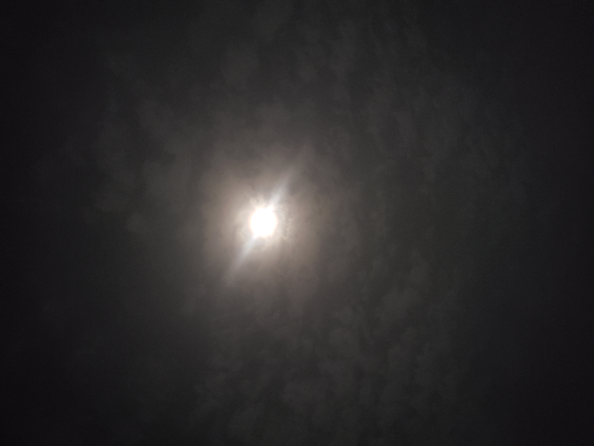 The image captures a celestial light source, most likely the moon, shining through an overcast night sky over Adankolo, Nigeria. The central light is intensely bright, appearing as a luminous white orb with a distinct starburst effect, indicating its powerful glow penetrating through atmospheric haze or clouds.
The sky is dark, predominantly black and deep grey, filled with varying densities of clouds. These clouds are illuminated from behind by the central light source, creating a soft, diffused halo around it and partially obscuring its exact shape. The cloud patterns show subtle variations in texture and density across the frame. The overall scene suggests a cloudy night with the moon, or another bright celestial body, providing the sole significant light. No other objects, people, or specific ground features are visible. There is no discernible activity taking place, as the image is a static capture of the night sky. No text is visible in the image.