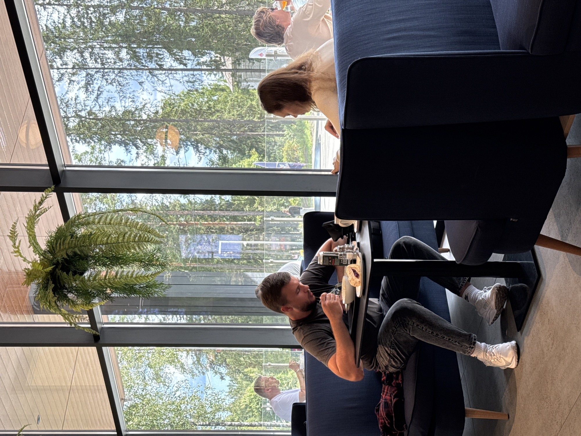 The image depicts the interior of a modern cafe with large windows offering a view of a forest. In the foreground, a man is seated at a table, eating from a bowl with a fork and a knife, with a slice of cake on a plate beside him. A can and a salt and pepper shaker are also on the table. He is wearing a dark t-shirt, grey jeans, white socks, and white athletic shoes. To his left, a red and black plaid shirt is draped over the dark blue booth seating.
In the background, through the window, a woman with long brown hair is seated at another table, facing away from the camera, her hands resting on the table. To her right, a man with blond hair and glasses is also seated, with a glass of amber liquid in front of him. Further in the background, through the window, other patrons are visible, along with trees and a building. A hanging fern adds a touch of greenery to the cafe's interior.
The cafe appears to be in Lakhdenpokhya, Russia, given the context provided. The overall atmosphere is casual and relaxed.