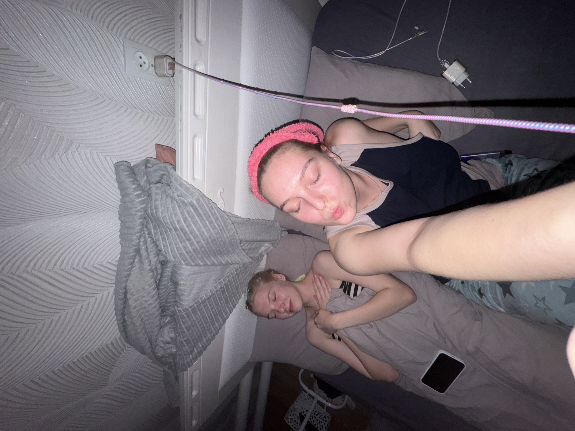 Two young women are in bed. The woman on the left is wearing a striped bandeau top and is under a gray blanket with her hands clasped over her chest. The woman on the right is wearing a pink headband and a dark tank top with a light-colored undershirt, and she is puckering her lips as if for a kiss. A phone is visible on the blanket near the woman on the left. There is a power outlet on the wall with a charger plugged into it, and a cable runs from the charger towards the women. The ceiling above them has a textured pattern, and a folded gray blanket is draped over a surface above the bed. The setting appears to be a bedroom in Zhigalki, Russia.