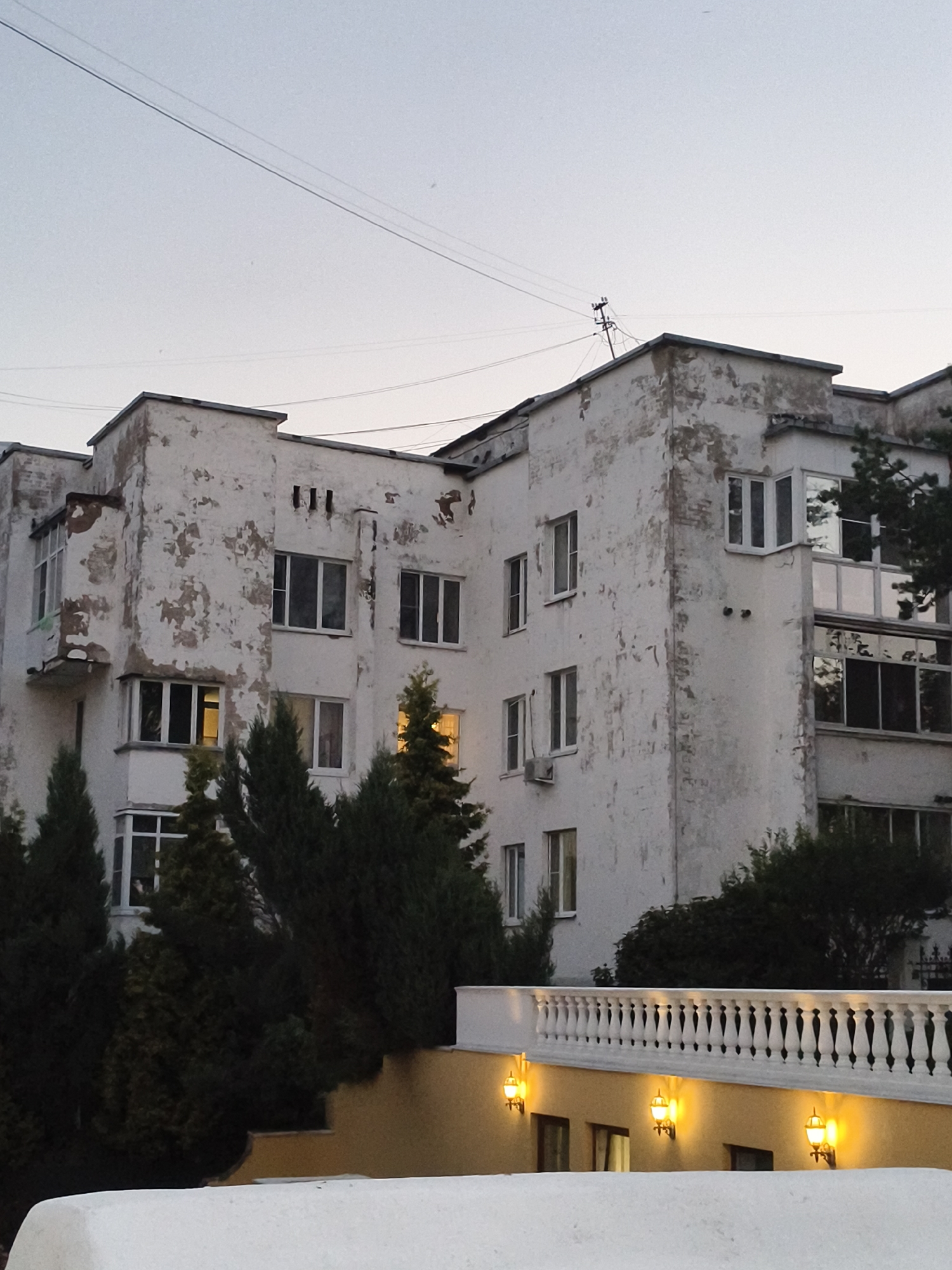 The image depicts a multi-story building with weathered white facade in Yaroslavl, Russia. The building exhibits signs of age, with peeling paint and visible brickwork in several areas. It features numerous windows, some of which are illuminated from within, suggesting occupancy. The architecture appears to be somewhat dated, possibly from the Soviet era, with distinct architectural elements like the stepped roofline and recessed balconies.
In front of the building, a line of evergreen trees, likely cypress or juniper, forms a dense green barrier. Beyond the trees, a yellow-painted wall is visible, topped with an ornate white balustrade. Several decorative outdoor lamps are mounted on the yellow wall, casting a warm glow. The sky above is overcast and pale, indicating either early morning or late evening light. Power lines are strung across the sky, connecting to a utility pole on the building's roof. The overall scene suggests an urban residential area undergoing some degree of decay, yet with attempts at beautification through lighting and landscaping. No people are visible in the image.