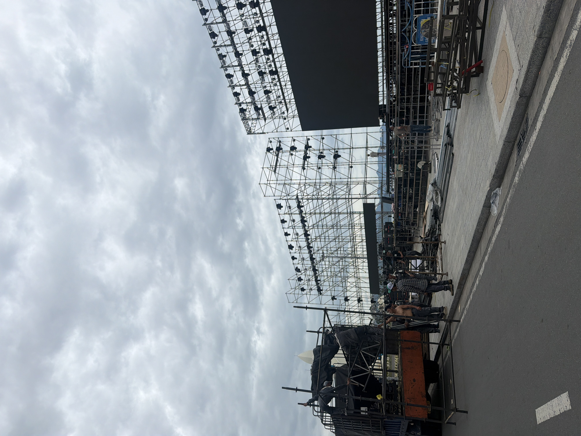The image depicts an outdoor scene in Nha Trang, Vietnam, where a large stage structure is under construction. The sky is overcast with heavy clouds. In the foreground, a truck with a black tarp-covered structure is parked on the left, with several individuals working on or around it. They are engaged in various tasks, appearing to be setting up or dismantling equipment. A group of men, some shirtless, are interacting near the truck. Further back, a massive scaffolding structure extends upwards, adorned with numerous spotlights. To the right of this scaffolding, a large, dark, rectangular screen, presumably a video display, is mounted on a similar metal framework. More scaffolding and lighting rigs are visible in the background. The ground is paved with concrete slabs and some areas of asphalt, suggesting a public or event space. The overall atmosphere is one of active preparation for a large-scale event, with construction workers diligently carrying out their duties.