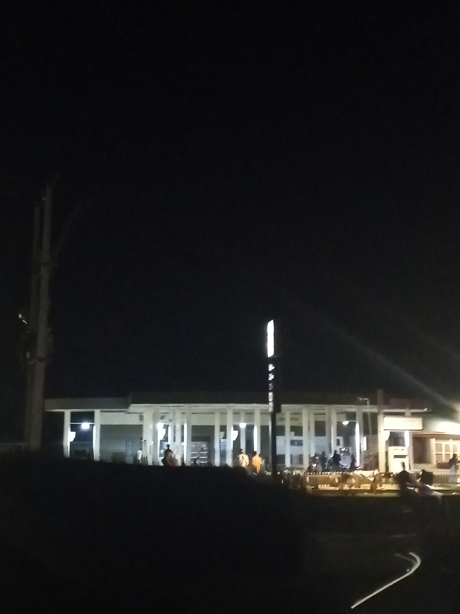 At night in Lokoja, Nigeria, a brightly lit, white-columned building stands, likely a commercial or public space. The scene is mostly dark. A utility pole rises on the left. Several people, appearing as silhouettes, are gathered in front of the building. A bright light pole stands to the right of center.