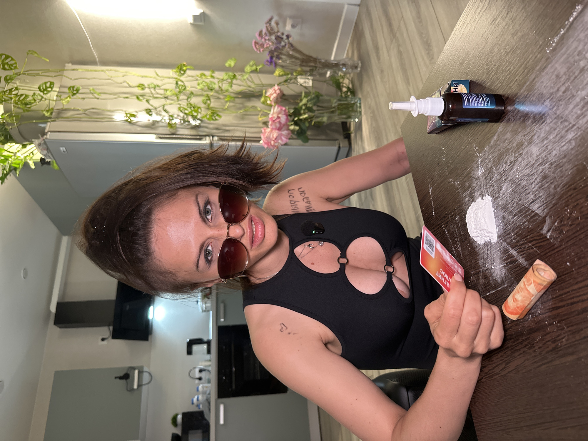 A woman wearing sunglasses and a black outfit with cutouts is sitting at a wooden table in what appears to be a kitchen. She is holding a credit card and looking at the camera. On the table in front of her, there is a pile of white powder, a rolled-up banknote, a small bottle with a dropper, and a pack of cigarettes. The background shows kitchen cabinets, a refrigerator, and plants. The overall lighting suggests an indoor environment.