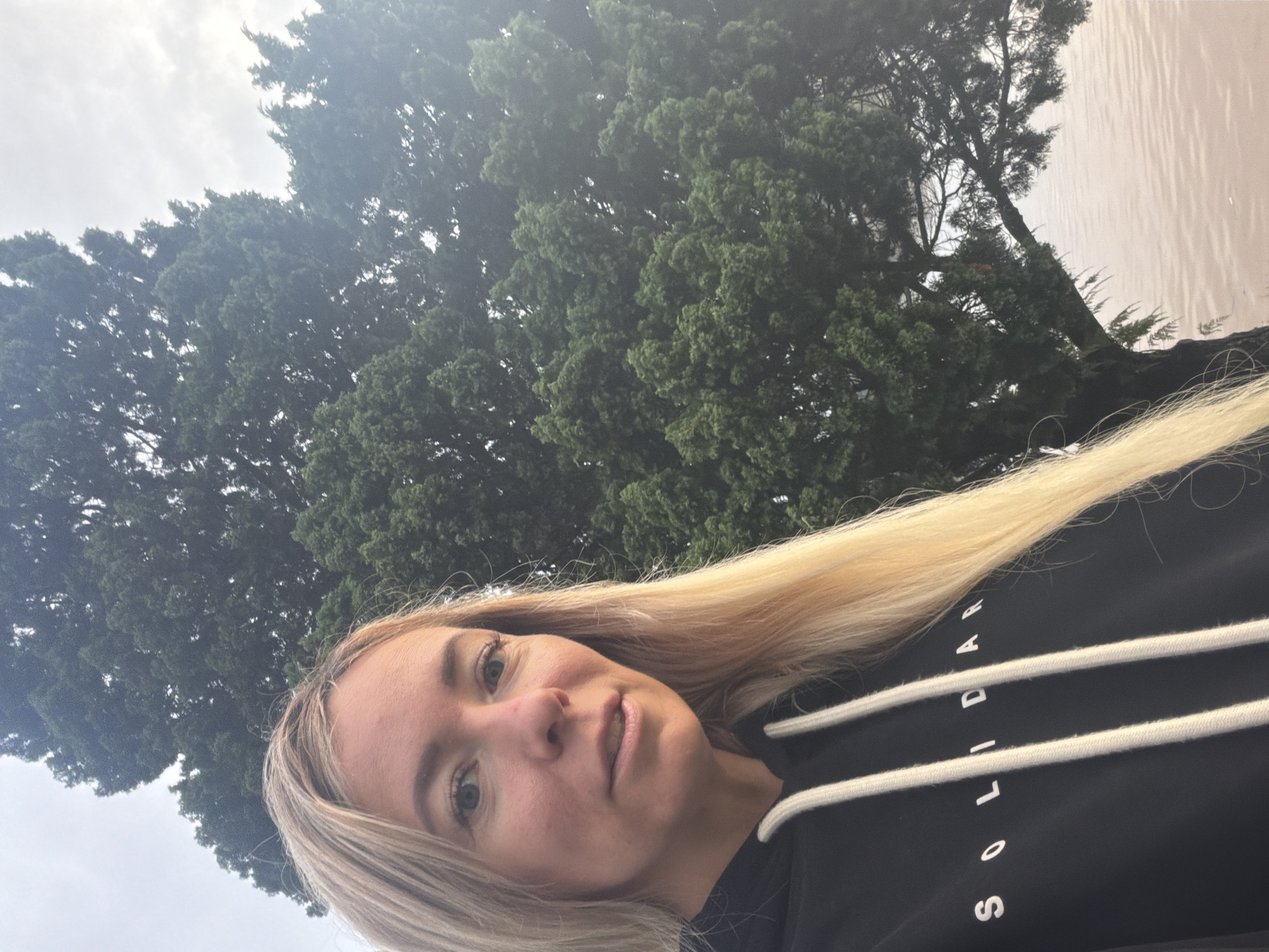 In a close-up selfie, a blonde woman with blue eyes and fair skin faces the camera. She wears a black hoodie with white drawstrings and the word "SOLIDAR" in white lettering across the chest. Her hair is long and blonde, falling over her right shoulder. Behind her, a large, dense green tree dominates the background, its branches reaching towards a pale, overcast sky. To the right of the tree, a body of murky, light-brown water is visible, reflecting the subdued light. The scene appears to be outdoors, with the natural elements of a tree and water suggesting a park or lakeside setting. The specific location in Da Lat, Vietnam, is not explicitly identifiable from the visual cues alone.