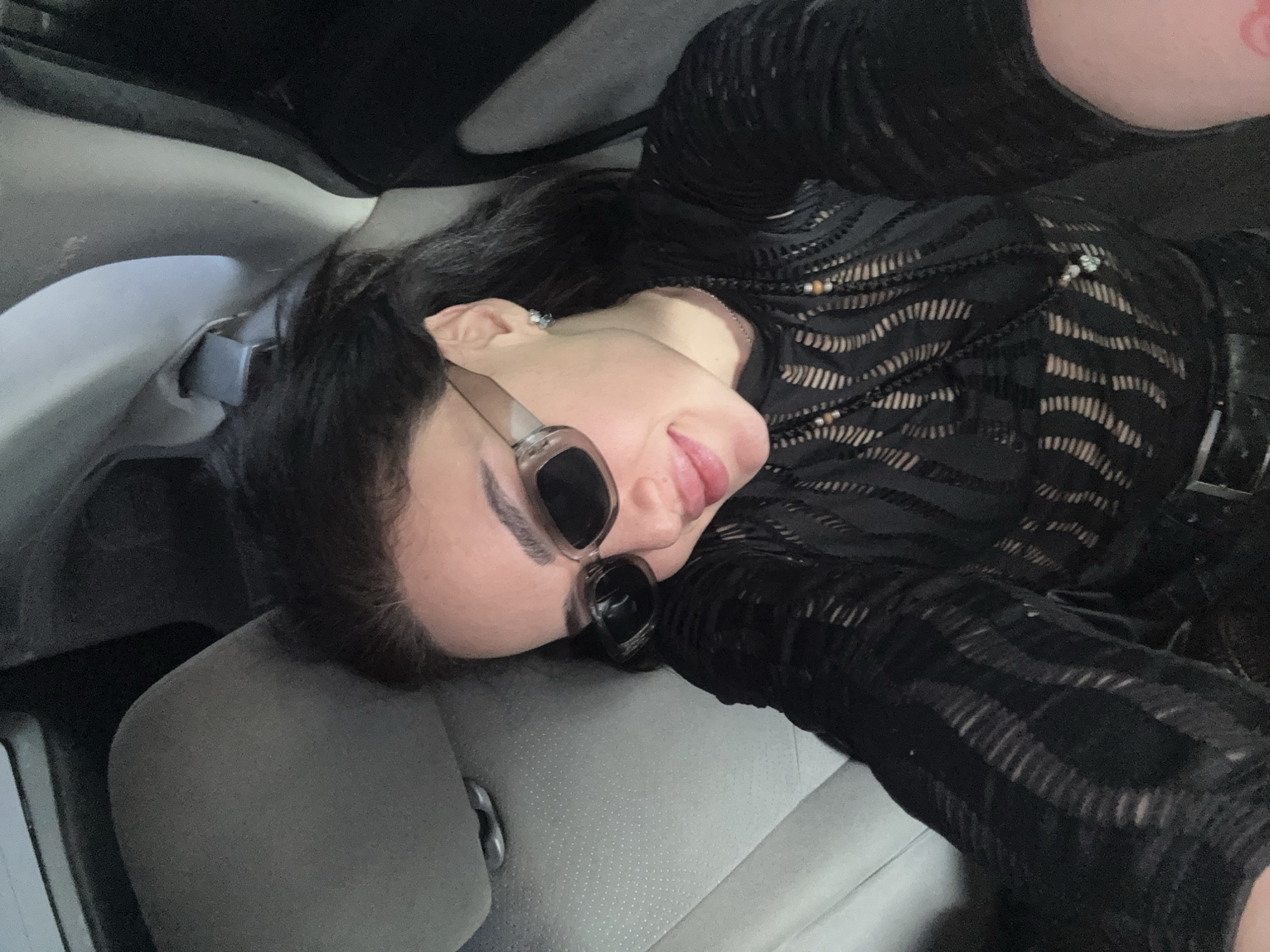 A woman wearing sunglasses and a patterned black top is reclining in a car. She appears to be taking a selfie. The location is indiscernible from the provided image.