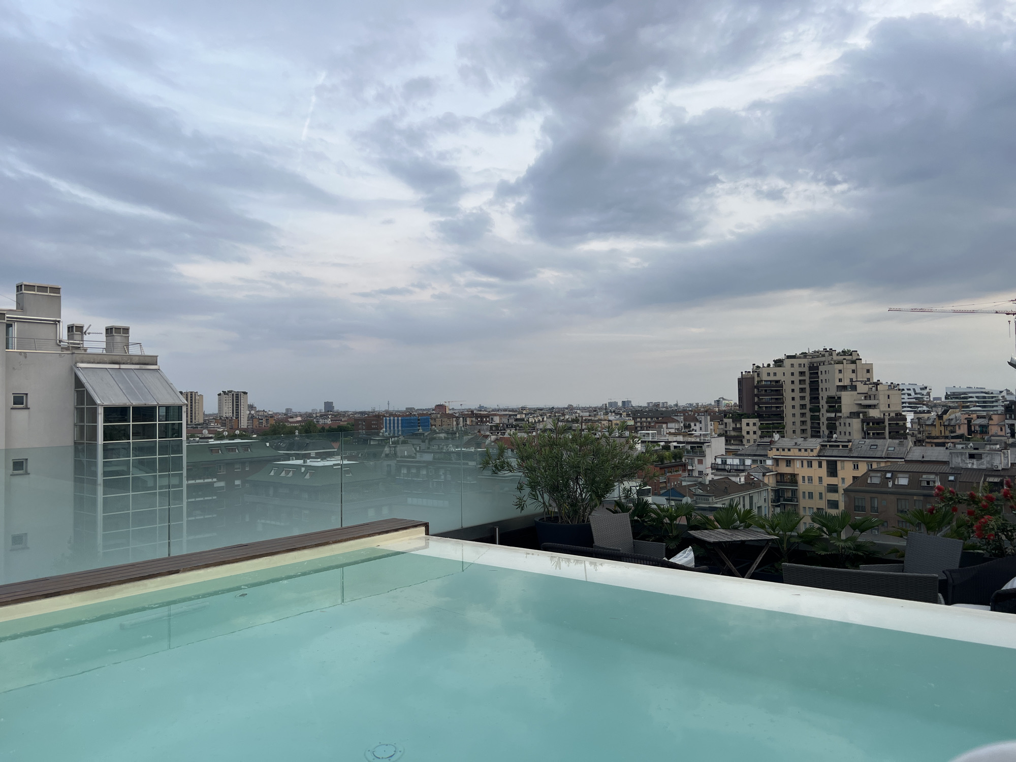This image captures a wide-angle, elevated view from a rooftop, overlooking a vast urban landscape under a cloudy sky. In the foreground, a rectangular swimming pool or hot tub with light blue water occupies the lower half of the frame, its surface reflecting the overcast sky. A dark wooden trim lines the nearest edge of the pool.
Separating the pool area from the city beyond is a clear glass barrier, through which the urban environment is visible. To the left, a modern building with a distinctive glass-paneled section and flat grey roof, complete with vents or chimneys, stands prominently. On the right side of the pool, a sophisticated rooftop terrace is visible, featuring a variety of lush green plants and small trees in pots, including palm-like foliage and some with red blossoms. Outdoor furniture, consisting of dark wicker chairs and a small wooden table, is arranged on this terrace.
The background reveals an extensive cityscape, characteristic of Milan, Italy. Numerous buildings of varying heights and architectural styles stretch toward the horizon. Taller, contemporary apartment blocks intermingle with older, multi-story buildings, many featuring pitched roofs and a mix of residential and commercial structures. A faint red construction crane is visible on the far right horizon, indicating ongoing development.
The sky is heavily overcast with layers of grey clouds, suggesting a cloudy day. No people or visible text are present in the image. The scene conveys a sense of calm, urban luxury, and expansive city life from a privileged vantage point.