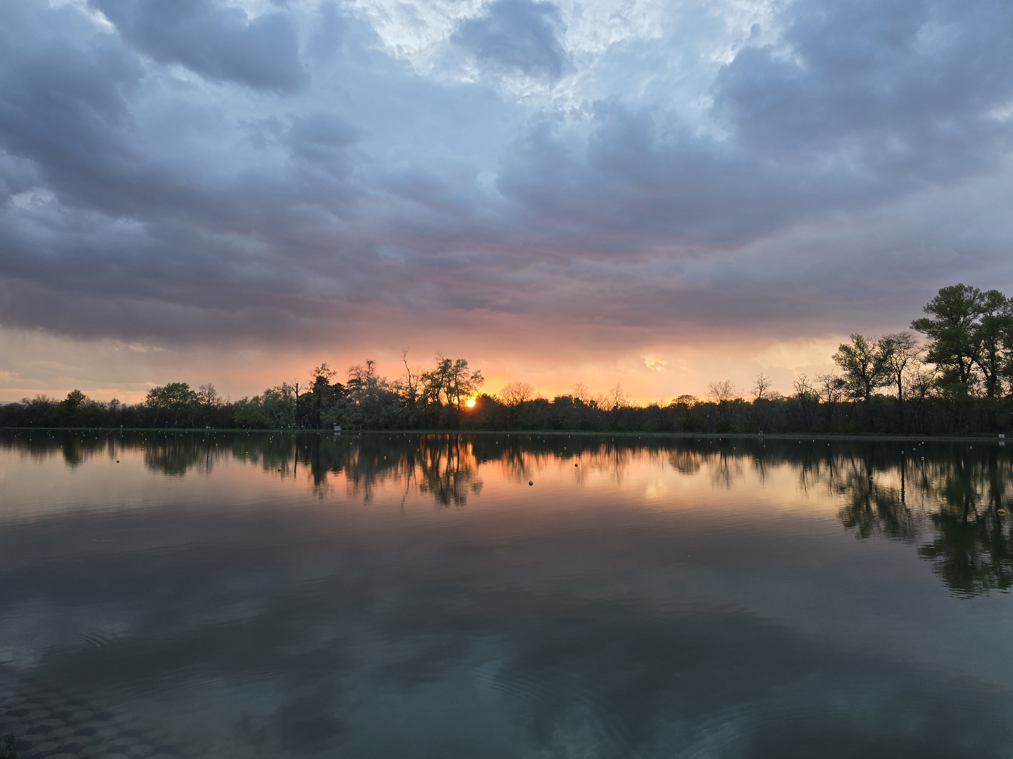 The image captures a serene sunset scene over a body of water, likely a lake or pond, in Unknown, Unknown. The sky is filled with dramatic, layered clouds, ranging from dark blues and grays in the upper portion to vibrant oranges and pinks near the horizon where the sun is setting. The sun is partially obscured by silhouetted trees lining the opposite shore, casting a warm glow. These trees are reflected in the calm water, creating a mirrored effect. The water itself transitions from a dark hue in the foreground to a lighter, reflective surface that mirrors the sunset colors.