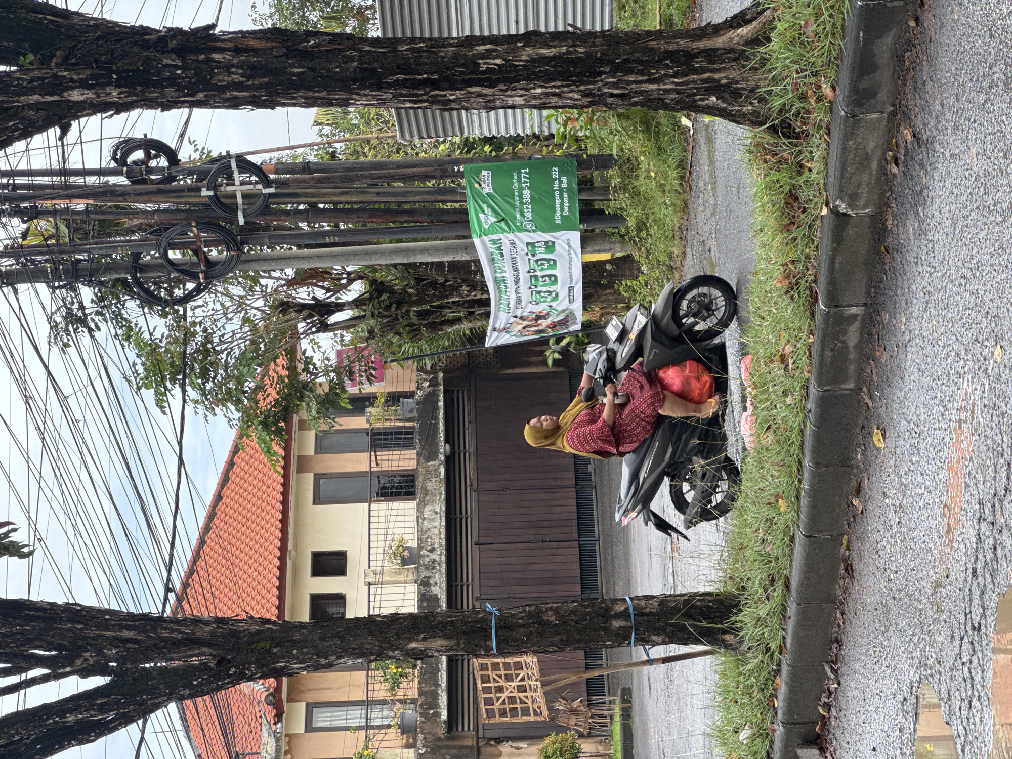 The image depicts a street scene in Kuta Selatan, Indonesia. A woman wearing a yellow hijab and a red patterned dress is seated on a black motorbike parked on the side of the road. She is holding a mobile phone. A banner is affixed to a utility pole that features the phrases "Qurban Durian" and "Qurban Menguatkan Sesama". A green portion of the banner shows "Jl Diponegoro No. 222 Denpasar - Bali" and a phone number. The buildings have light-colored walls with red tiled roofs. Wires run across the sky in front of the buildings. A tree trunk with blue ties is visible in the foreground. The road appears wet.