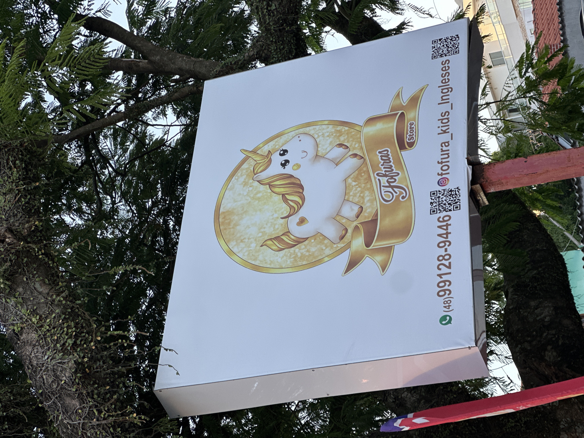 A white square sign displaying a cartoon unicorn and the name "Fofuras Store" in decorative script is mounted outside a business. The sign also features a WhatsApp number, Instagram handle "fofura_kids_Ingleses," and two QR codes. The sign appears to be attached to a tree and is partially obscured by branches. The image was taken in Florianópolis, Brazil.