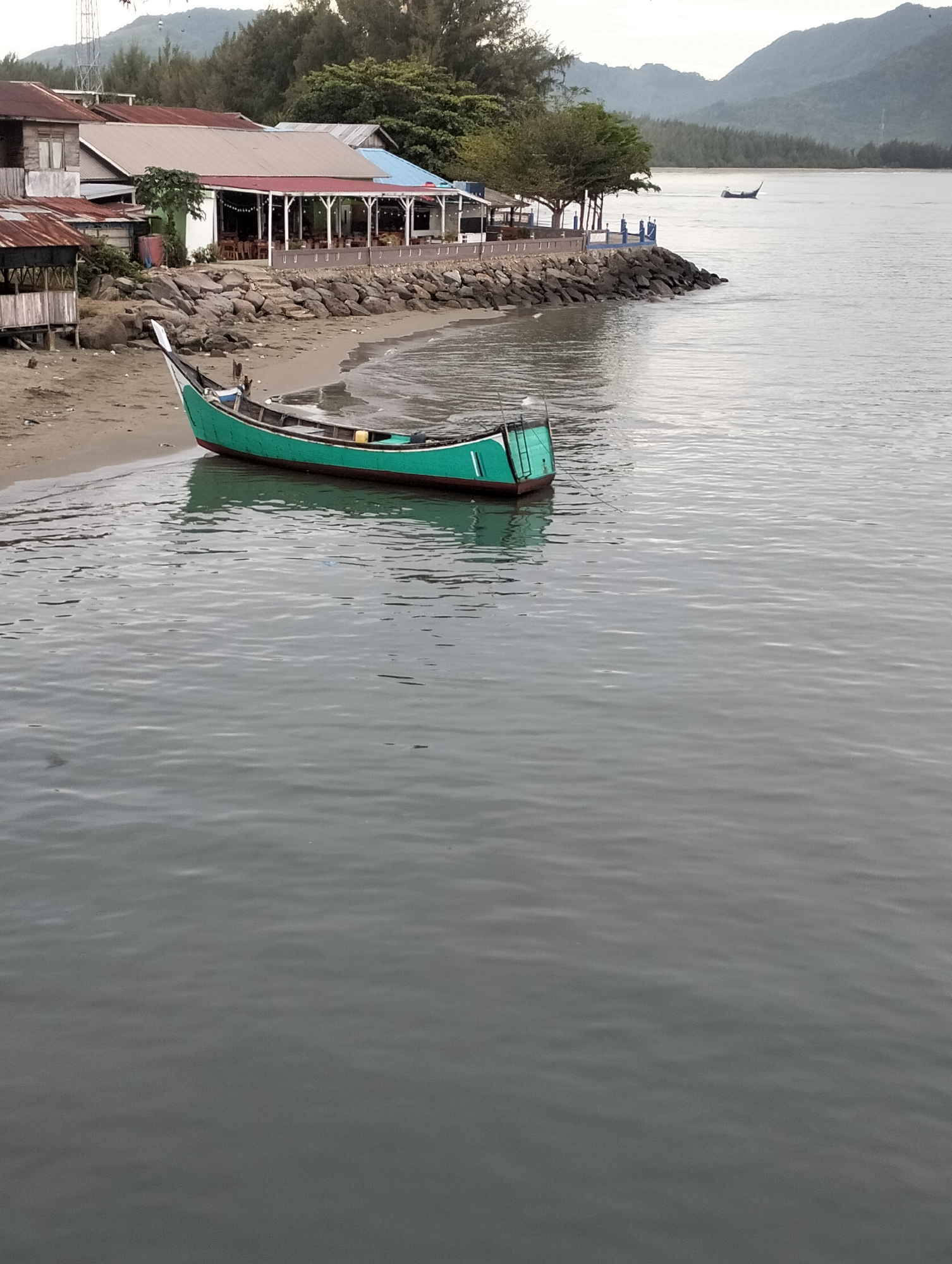 The image depicts a coastal scene in Banda Aceh, Indonesia, featuring a green wooden boat partially beached on a sandy shore. The boat is the primary subject, resting between the sand and the water. Behind the beach, a structure with a red roof and open sides suggests a restaurant or public space. A rock barrier separates the building from the shoreline. In the background, tree-covered hills or mountains are visible across the water. The sea is relatively calm, with a few ripples on the surface. Another boat is further out in the water in the distance. The lighting suggests either early morning or late afternoon.