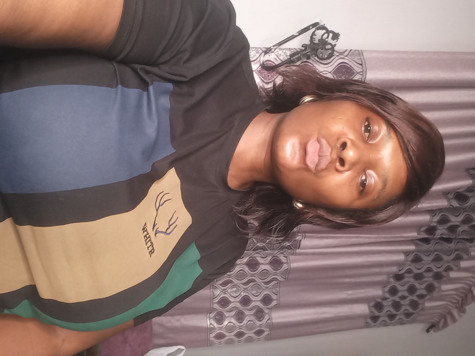 An adult woman is seen from the shoulders up, slightly off-center, facing the camera with a pursed-lips expression. She wears a black t-shirt with horizontal stripes in blue, beige with the word "WHITE" and deer antlers printed on it, and green. She has shoulder-length brown hair, and is wearing gold hoop earrings. The background is a patterned fabric. The location is Jos, Nigeria.