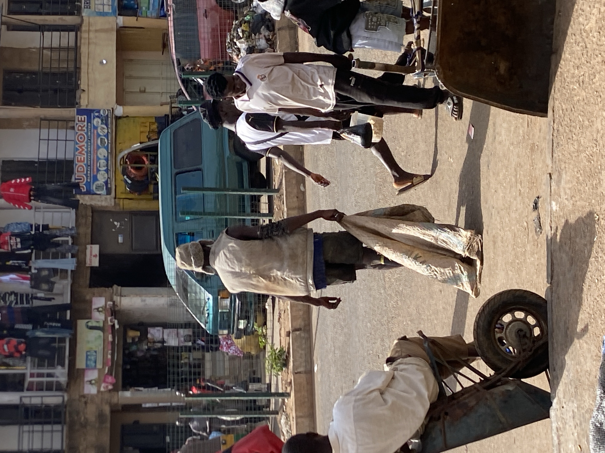 The image depicts a street scene in Bukuru, Nigeria. Three men walk along a paved area in the foreground; two wear white shirts and dark pants, and the third man is further back wearing a light vest and dark shorts. A man pushes a wheelbarrow. A light blue van is parked along the edge of the street in the background. Buildings are visible on the left side of the frame, and signage with the word "JUDEMORE" is displayed on one of the buildings. Clothes hang on racks in front of the buildings. The environment appears to be a busy urban area.