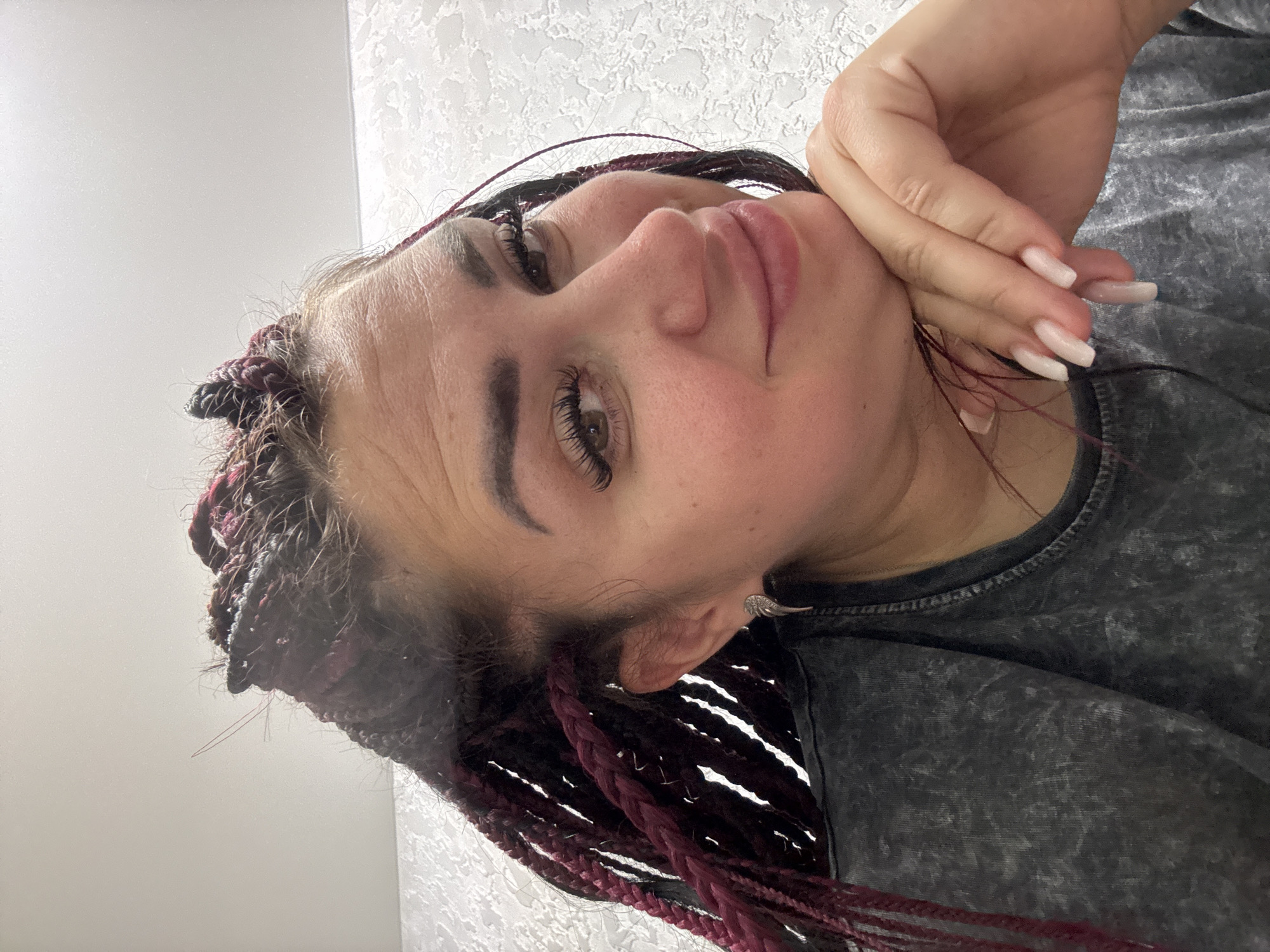 A woman is shown in a close-up selfie. She has dark braided hair with burgundy streaks and is wearing a grey t-shirt. Her right hand is resting on her chin and she is smiling slightly. The background appears to be a textured wall. The image was taken in the city of Tolyatti, Russia.