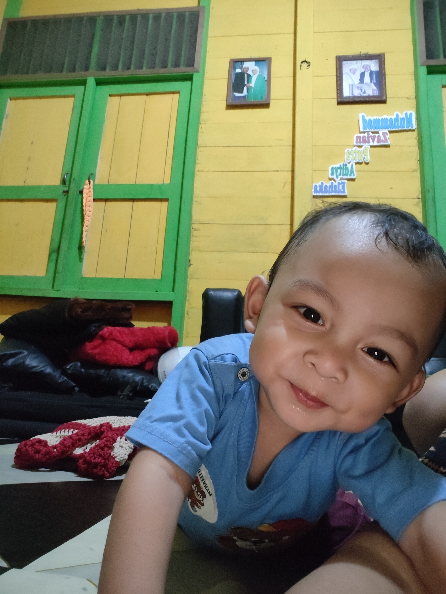 The image captures a close-up of a young child, possibly a toddler, smiling into the camera. The child is wearing a blue t-shirt. The background features a yellow wooden wall with green doors. There are framed pictures on the wall, and some cut-out letters arranged in a staircase pattern. A stack of pillows and blankets is visible to the left, and a crocheted blanket is in the foreground. The setting appears to be an indoor residential space, likely a home in Tanah Grogot, Indonesia, suggested by the style of the doors, wall paneling, and the text on the wall which may be in the local language. The lighting suggests the photo was taken indoors, possibly with natural light from a window or door.