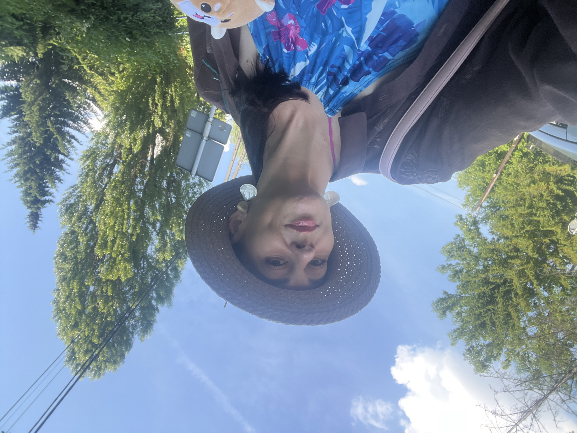 A woman wearing a wide-brimmed hat, sunglasses, and a blue floral halter top, is holding a stuffed animal. She is wearing a brown jacket and a light-colored purse is slung over her shoulder. The woman is outdoors with trees and a blue sky with clouds in the background. A traffic sign is visible to her right.