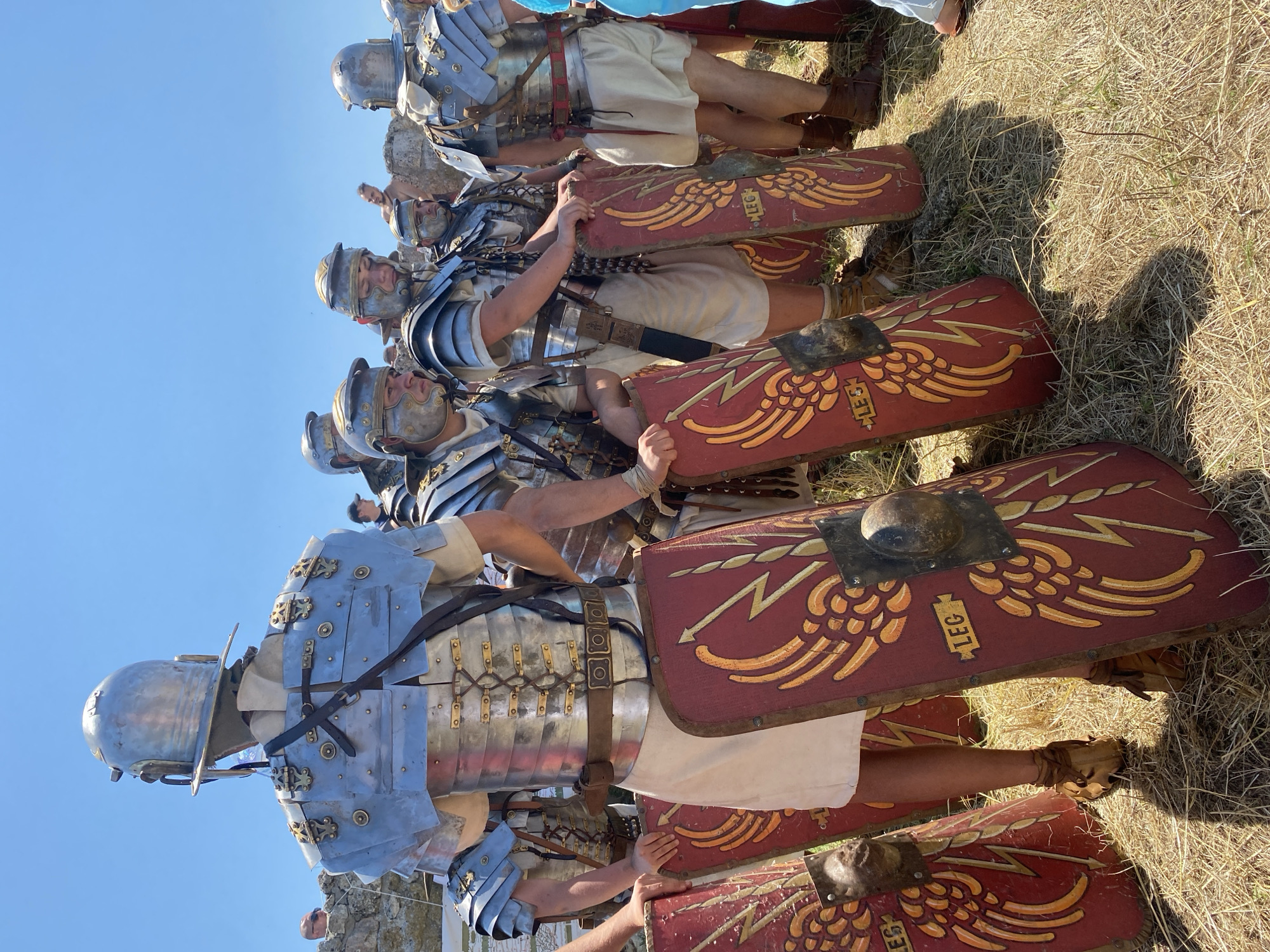 The image depicts a historical reenactment in Gigen, Bulgaria, featuring a group of individuals dressed as Roman legionaries under a clear, bright blue sky.
In the foreground, a male figure is observed from behind, wearing a polished metallic Roman helmet (galea) and a segmented plate armor (lorica segmentata) consisting of numerous silver-grey metallic bands secured with brass-colored rivets, hinges, and leather straps across the back and shoulders. A light-colored, possibly off-white, tunic is visible beneath the armor.
To the subject's right and further into the midground, several other male figures are similarly attired in metallic helmets with brass detailing, lorica segmentata, and light-colored tunics. One individual to the right wears a wide brown leather belt with square metallic fittings. The soldiers are standing on dry, sparse, yellowish-brown grass, casting distinct shadows from strong overhead sunlight.
Multiple rectangular Roman shields (scuta) are prominent, resting on the ground in front of the legionaries. These shields are predominantly red with a central yellow/gold winged lightning bolt motif, outlined in red, and feature the abbreviation "LEG" (for Legio) inscribed in a yellow rectangular cartouche. Each shield possesses a central, spherical metallic boss (umbo). Several individuals are seen holding these shields, their hands resting on the top edges. The legionaries wear brown, lace-up leather sandals (caligae).
In the background, fragmented stone structures and low walls, indicative of archaeological ruins, are visible under the expansive blue sky. Distant, partially visible modern observers are present, including one individual in sunglasses looking over a stone wall on the left, and another figure wearing a cap further right. A person in light blue attire is partially visible on the far right edge of the frame.