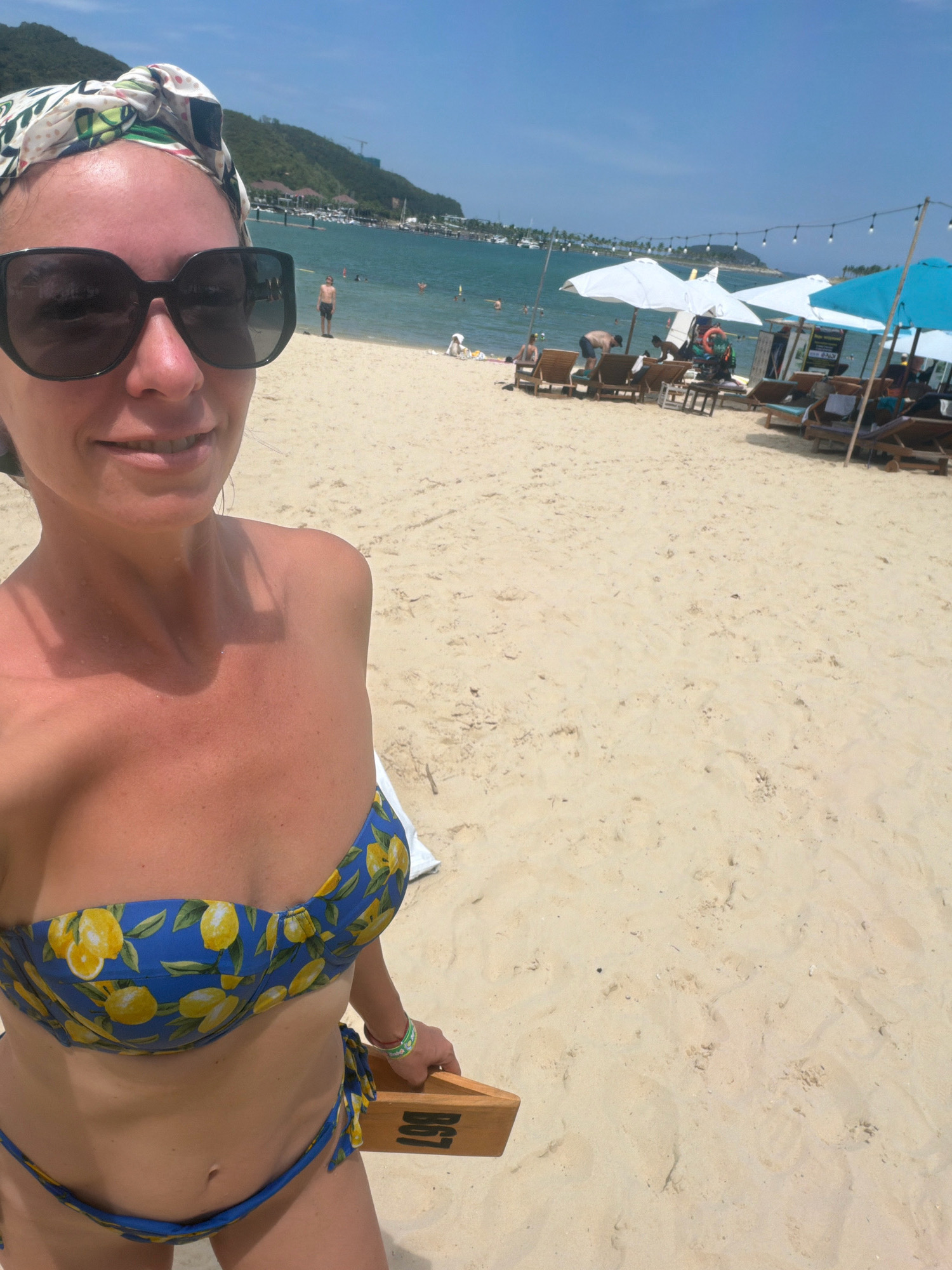 A woman in a blue and yellow lemon-print bikini and a white and green patterned headband poses for a selfie on a sandy beach in Nha Trang, Vietnam. She is wearing large black sunglasses. Behind her, the turquoise ocean stretches out, dotted with boats and swimmers. A line of white and blue beach umbrellas and lounge chairs are set up on the sand. Mountains with lush vegetation form the backdrop, and a clear blue sky completes the scene. The woman is holding a wooden plank with the word "BHT" printed on it.