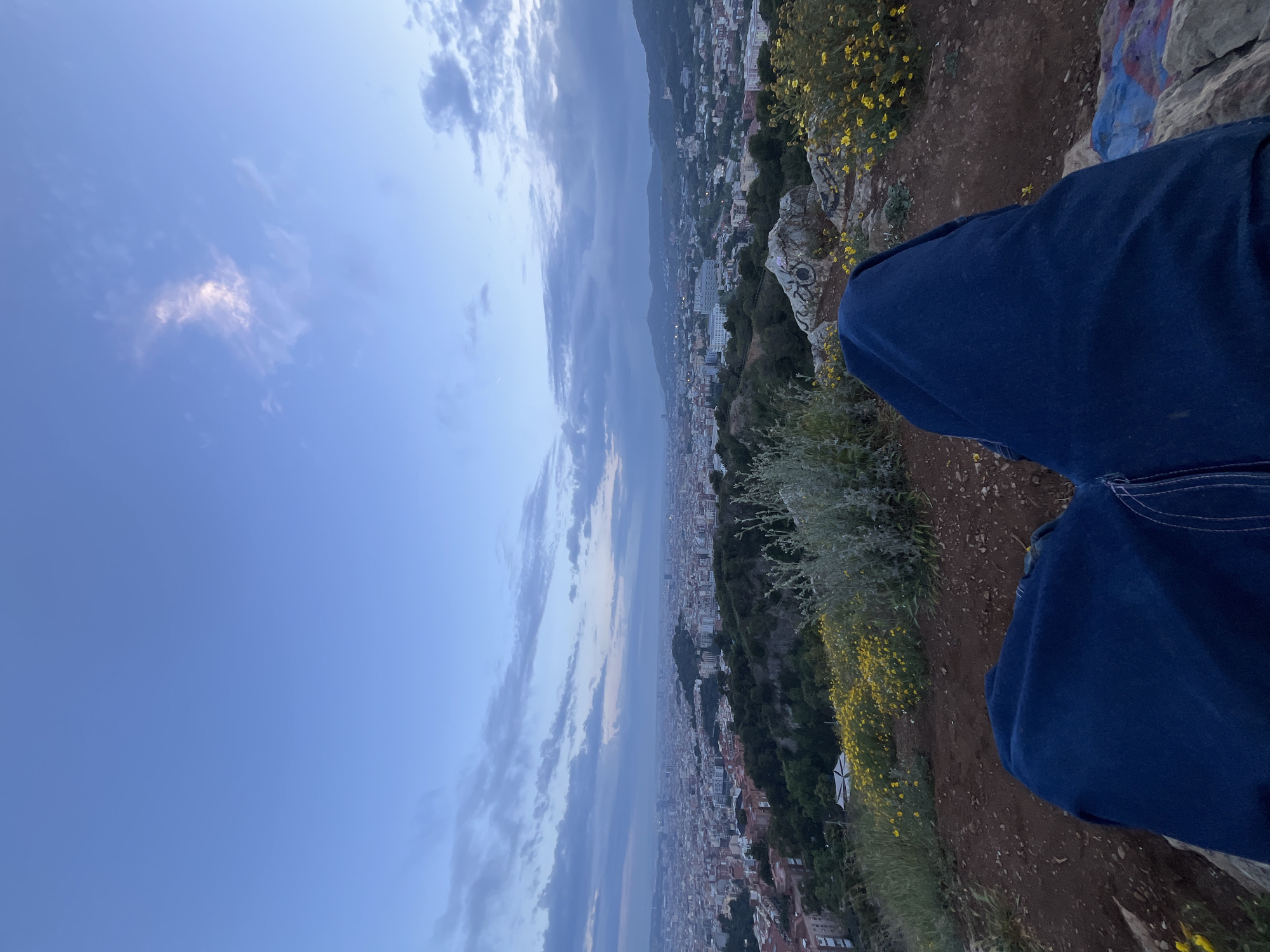A scenic view overlooks Barcelona, Spain, with a clear, cloudy sky above. In the foreground, blue denim-clad legs and a rocky, floral terrain occupy the right side of the frame. The cityscape stretches across the horizon, blending residential and commercial structures with patches of greenery and mountain slopes. The photo captures a perspective from a high vantage point, likely from a hilltop near the city, offering a panoramic view of the urban landscape under the evening sky.
