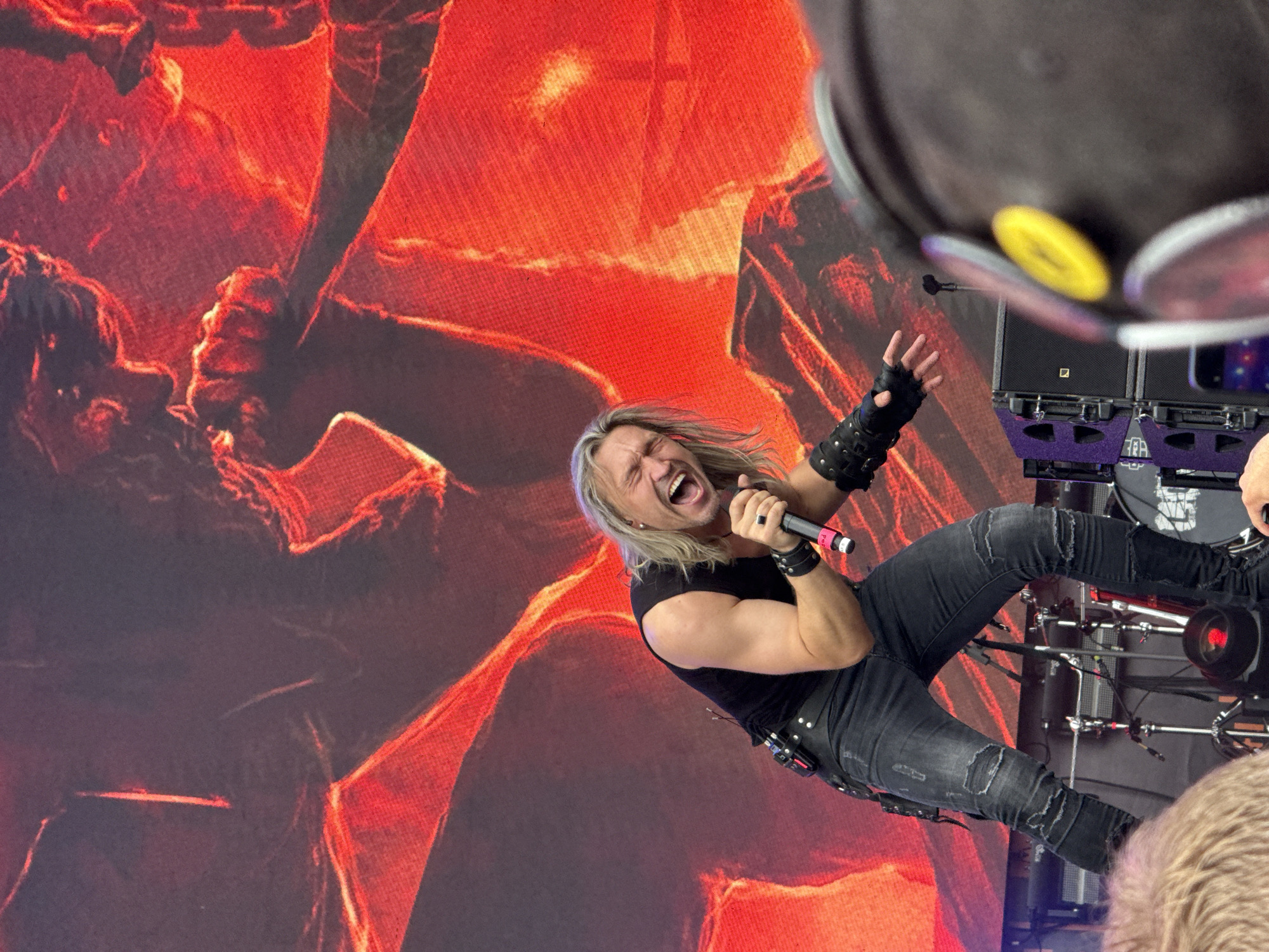 A male rock musician with long blonde hair, wearing a black sleeveless shirt, distressed black jeans, and black wristbands, is performing on stage. He is holding a microphone and appears to be shouting or singing loudly, with his mouth wide open and eyes closed. Behind him, a large screen displays a fiery, abstract red graphic. In the foreground, the heads of two audience members are visible, one with blonde hair and the other with dark hair wearing a black hat. The stage setup includes speaker cabinets and drum equipment. The setting appears to be an outdoor music festival.