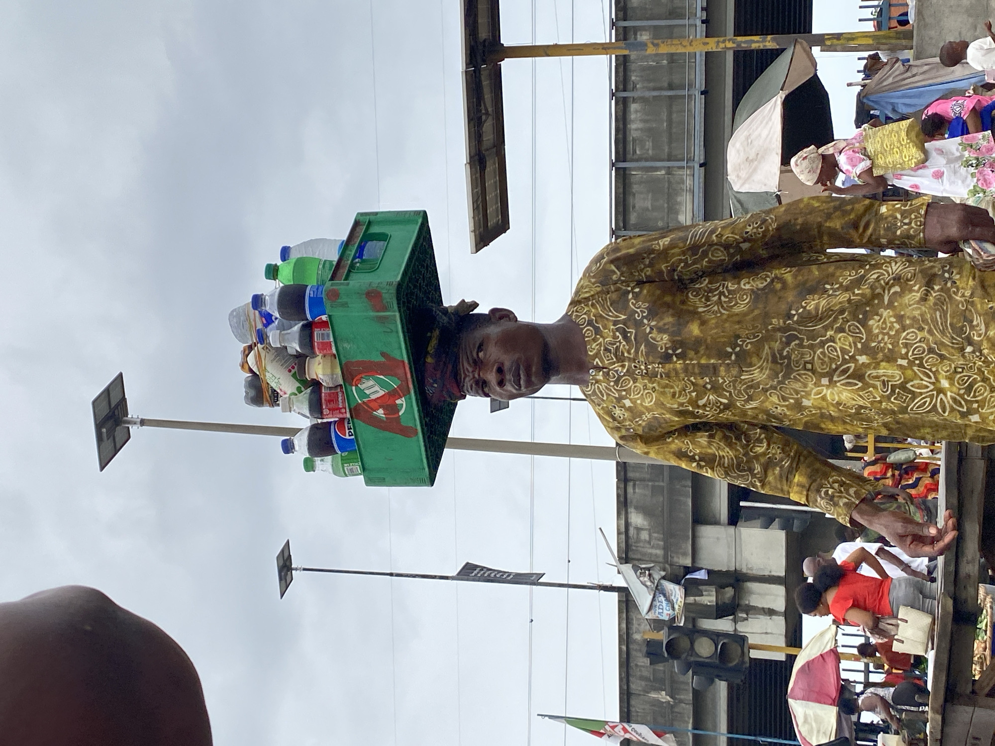 A street vendor in Lagos, Nigeria balances a green crate filled with assorted bottled drinks on his head. He wears a patterned, long-sleeved shirt. The background shows a bustling market scene with other people, stalls, traffic lights, solar-powered street lights, and overhead power lines.