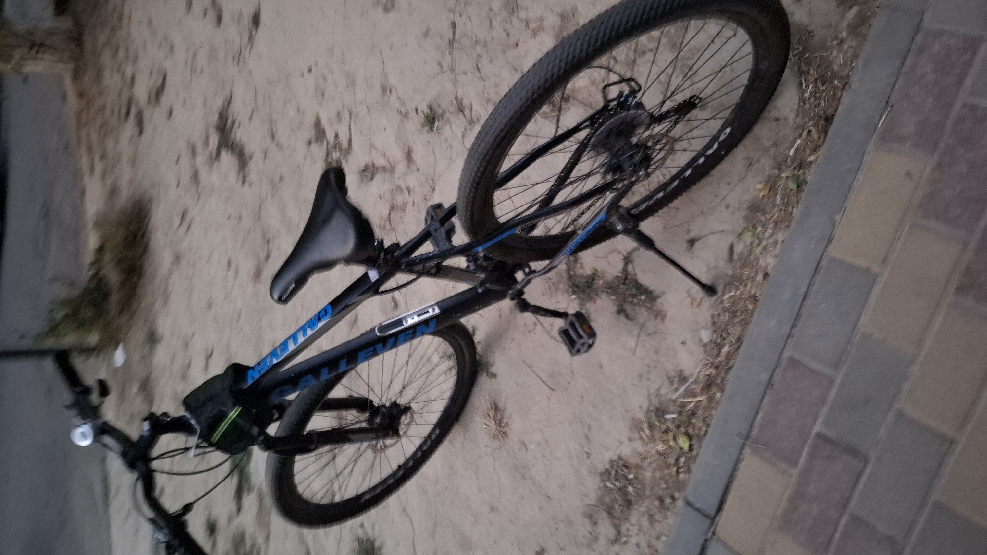 A black mountain bike is parked on a dirt path next to a paved walkway. The bike has blue accents on the frame and a black seat. The front wheel is slightly turned to the left, and the bike is supported by its kickstand. In the background, there are dry plants and a tree trunk. The image appears to have been taken in Shymkent, Kazakhstan.
